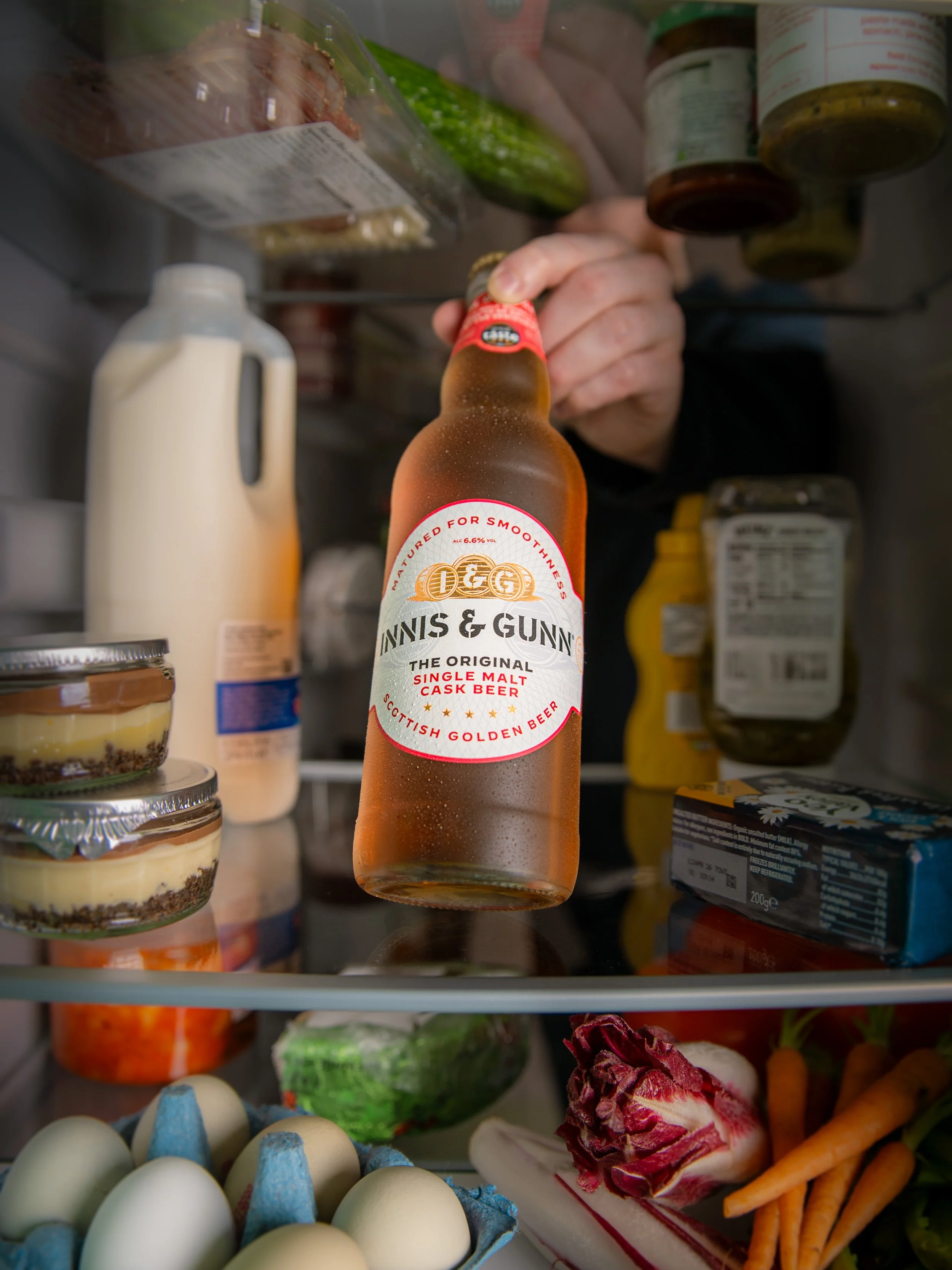 Inside a refrigerator, a hand is reaching to grab a bottle of Dennis & Glynne single malt Scottish golden beer. The fridge contains various food items, including eggs, fresh vegetables, a bottle of milk, containers of pre-made desserts, and jars of c