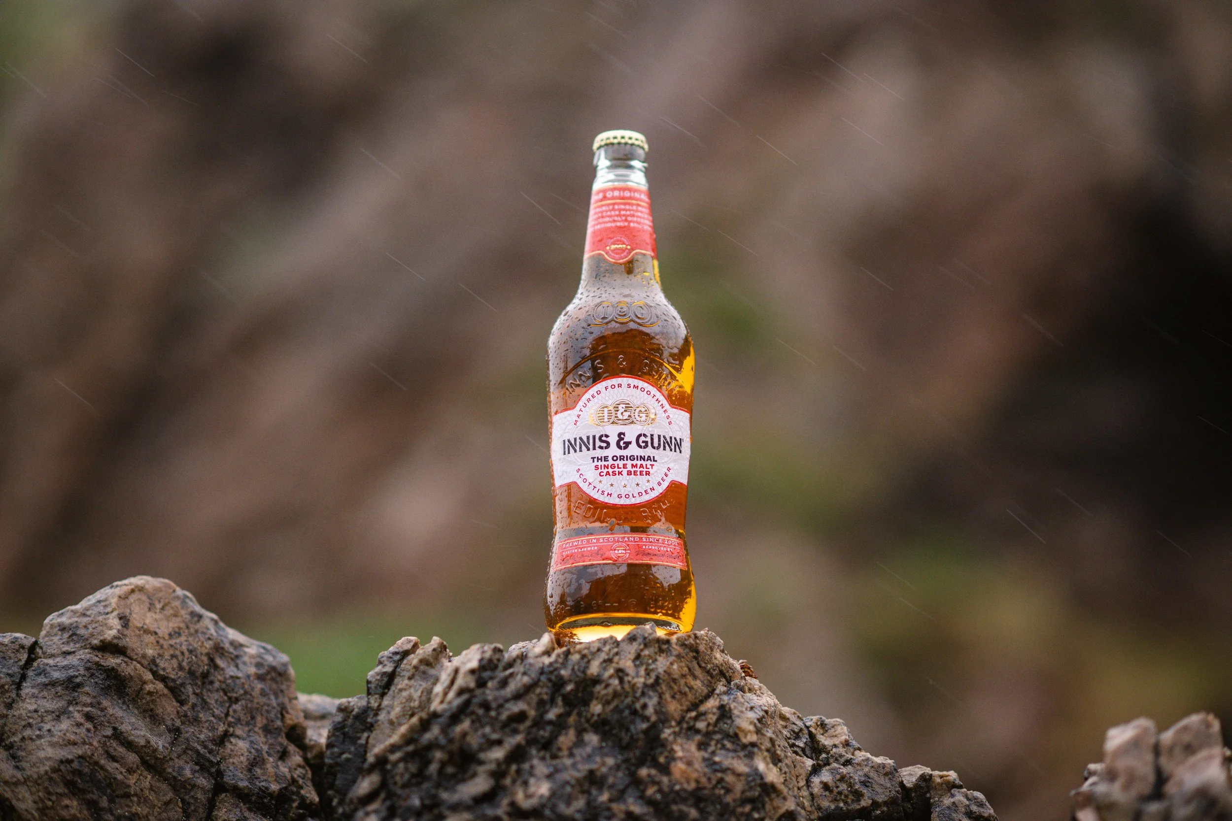 A bottle of Innis & Gunn original cask beer outdoors, placed on a rough rock surface in rainy weather.