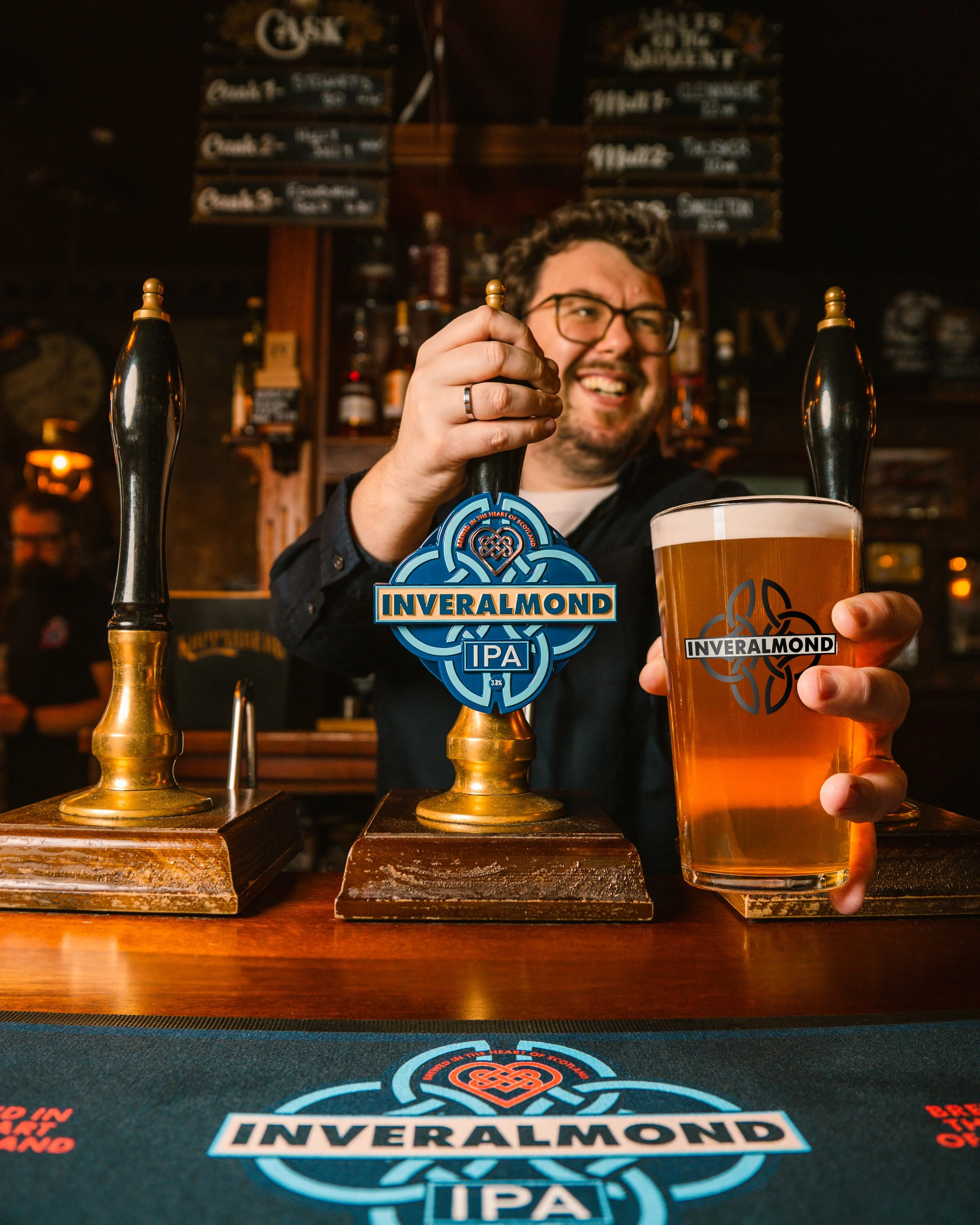 A bartender in a black shirt with glasses pours a beer from a tap labeled 'Inveralmond IPA' at a bar with a proud smile. There are two taps with black handles, one labeled 'Inveralmond' and the other with no visible label. The bar has a dark wooden b