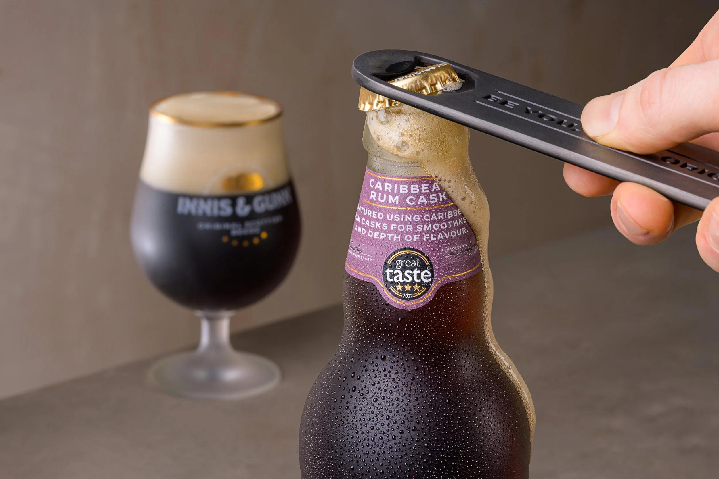 Person opening a bottle of Caribbean rum cask beer with a bottle opener, condensation on the bottle, in the background a glass of dark beer