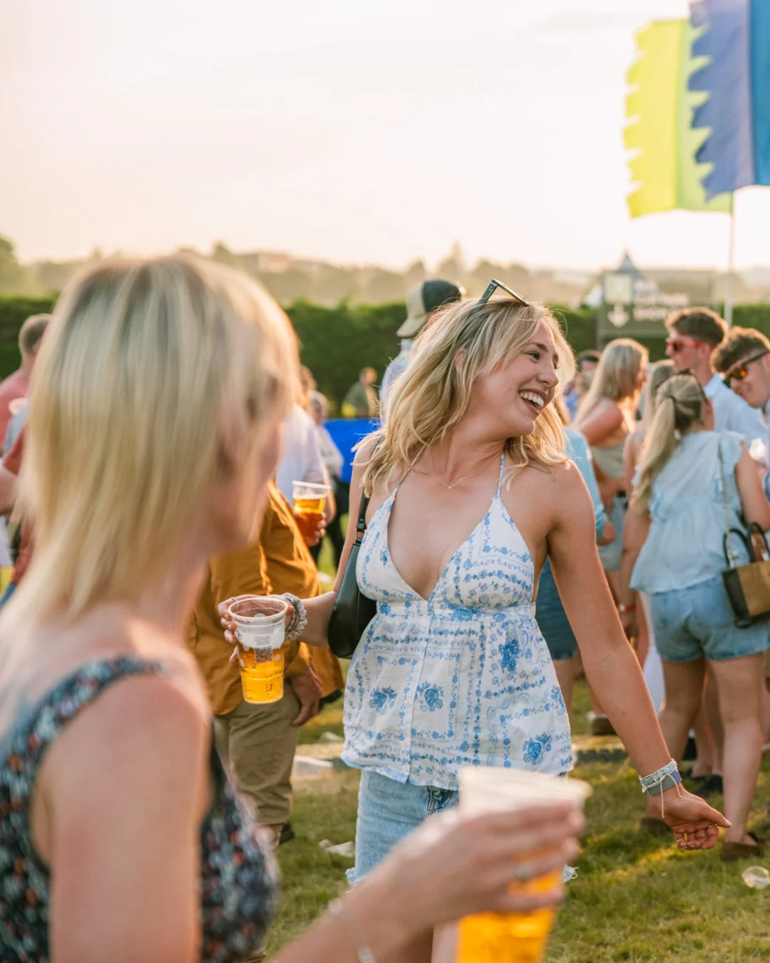 People enjoying a summer outdoor music festival, holding drinks and smiling, with a large colorful tent in the background.