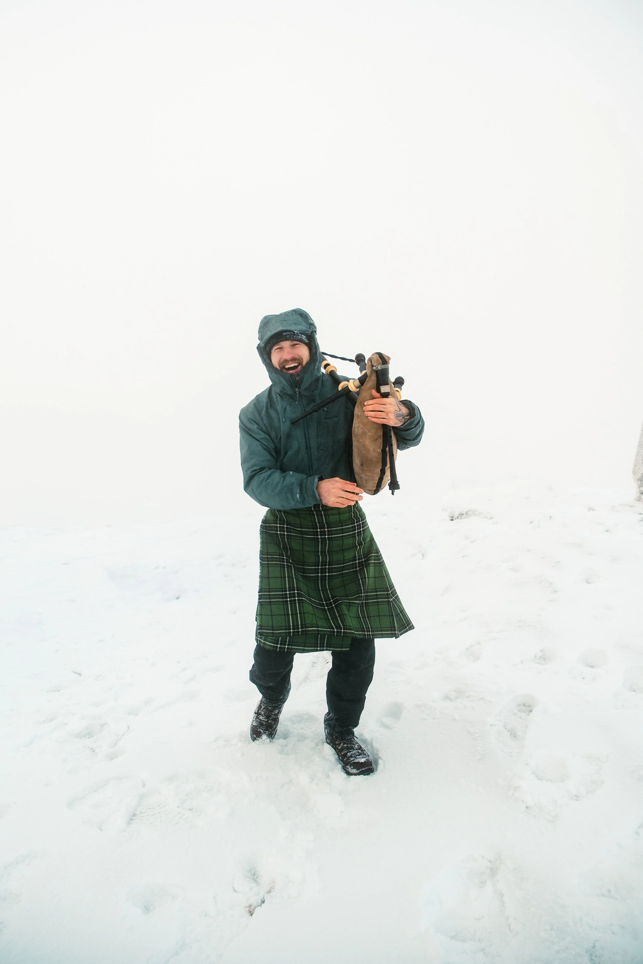 Person in winter gear, holding a bagpipe, standing in snow, smiling, with foggy white background.