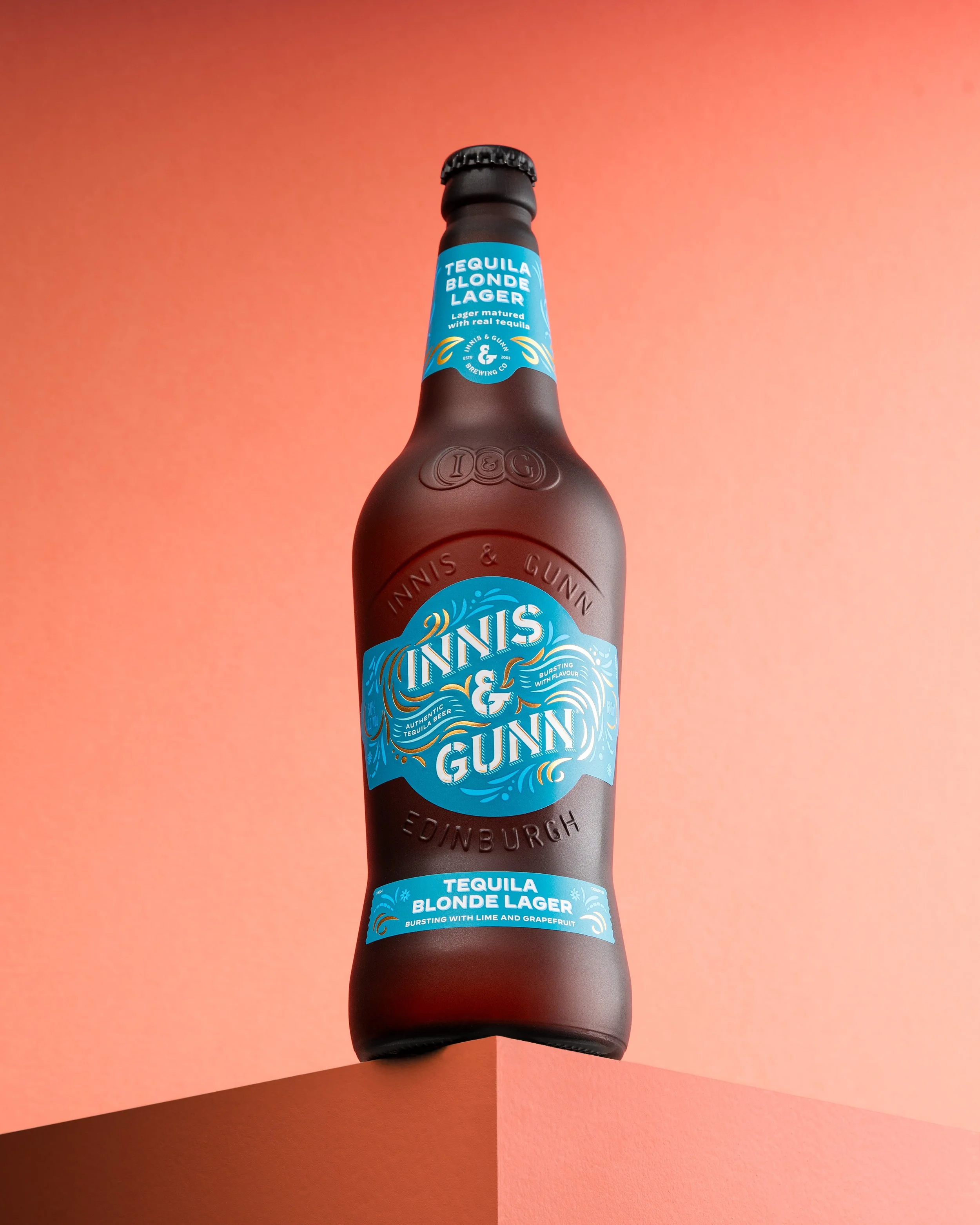 A dark brown glass bottle of Tanis & Gunn Tequila Blonde Lager, with blue and white label, placed on a pink surface against a gradient pink background.