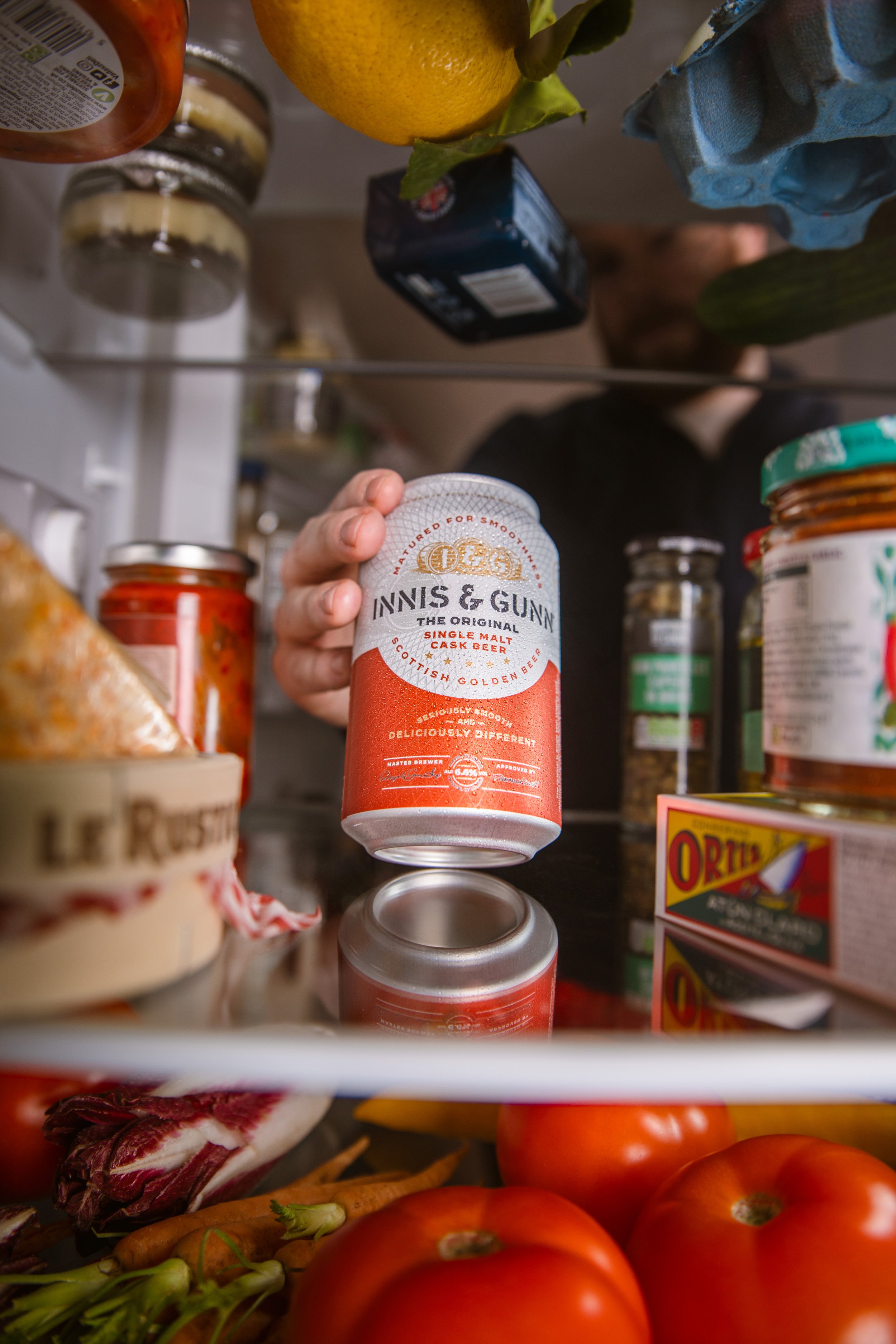 Inside a refrigerator, a person's hand holds a can of Inniskillin beer. The fridge contains various food items, including tomatoes, carrots, cheese, and condiments.