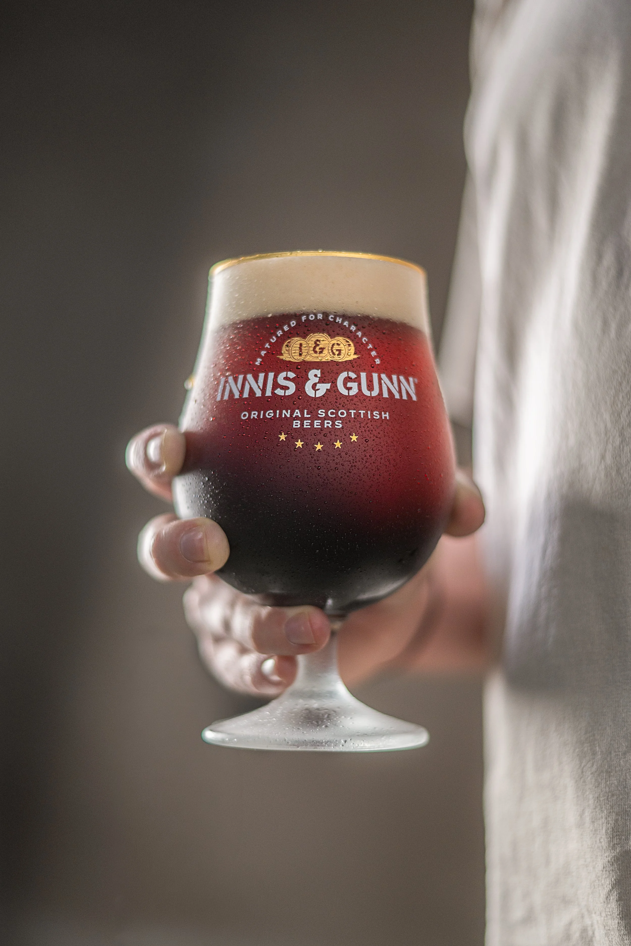 A person holding a glass of dark Scottish ale with foam head, branded with 'Innis & Gunn' logo, condensation on the glass, seen against a neutral background.