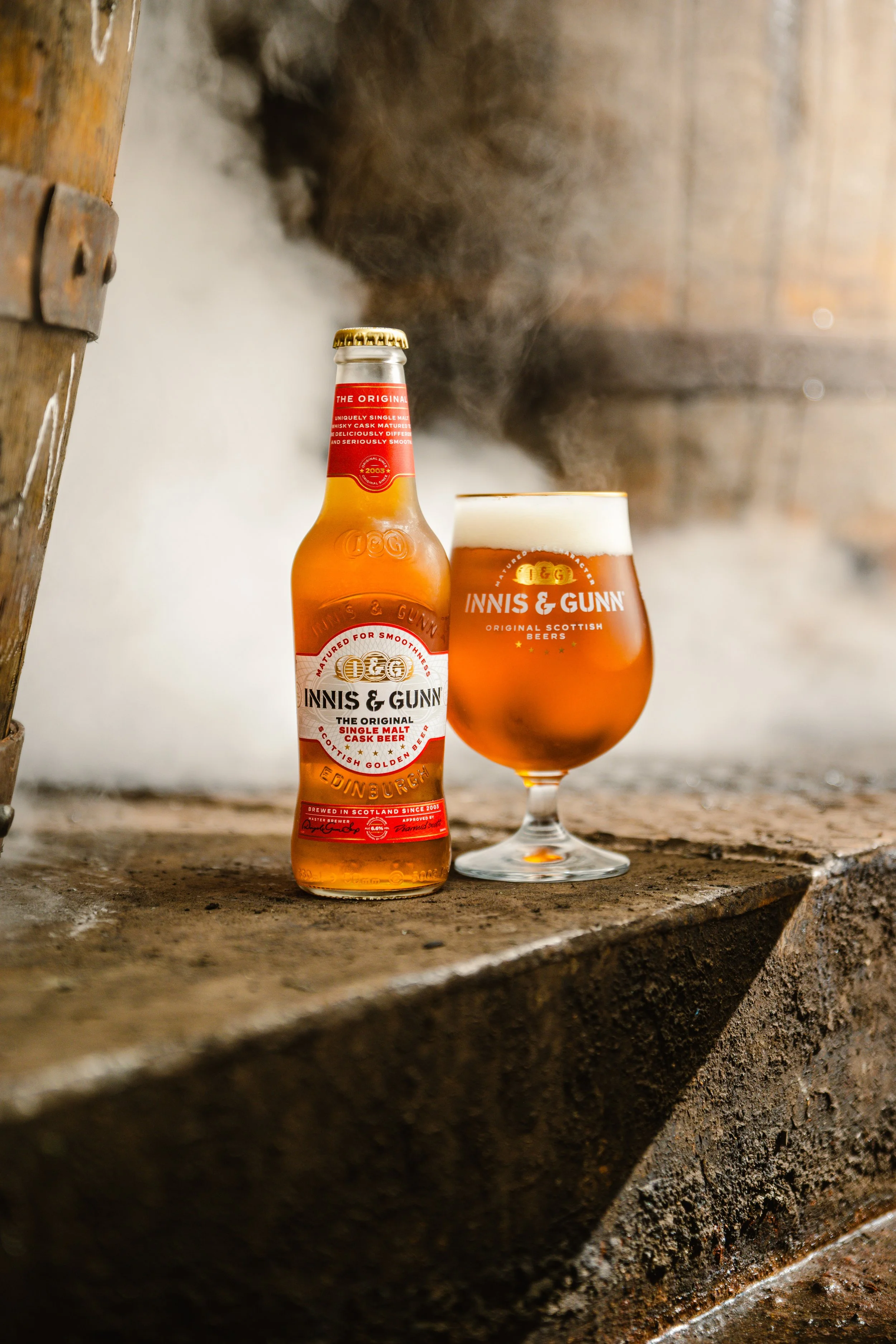A bottle of Innis & Gunn Scottish single malt cask beer and a filled beer glass on a rustic wooden surface with steam rising in the background.
