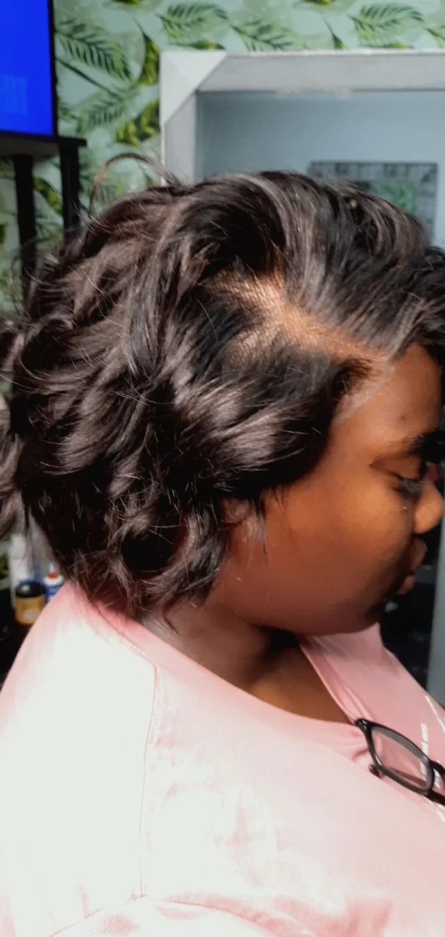 Side view of a woman with short, wavy black hair, wearing a pink shirt and glasses hanging from her collar, in a salon or similar setting.