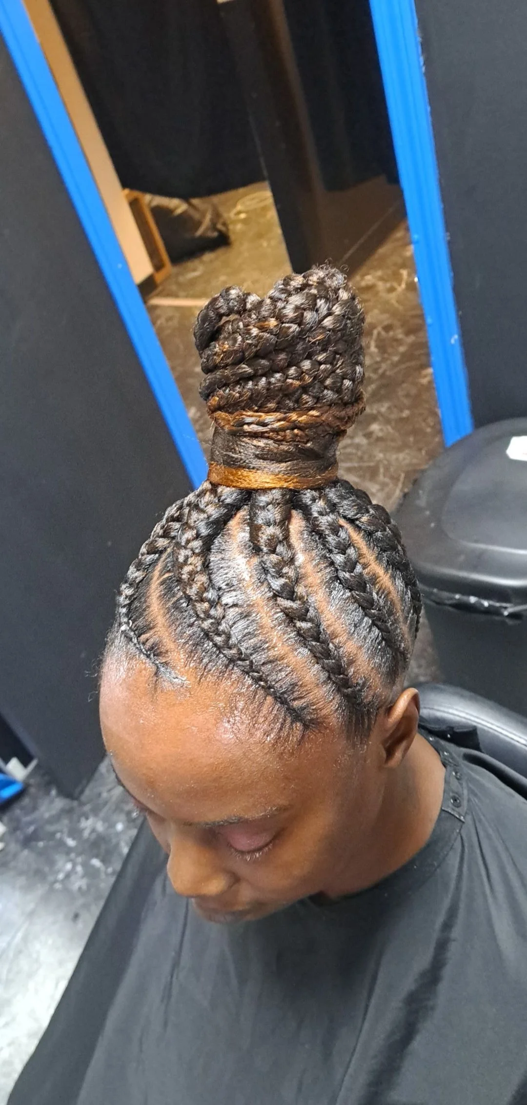 A woman with intricately styled braids in her hair, with the top braided into a large bun twisted at the crown of her head, in a salon setting.