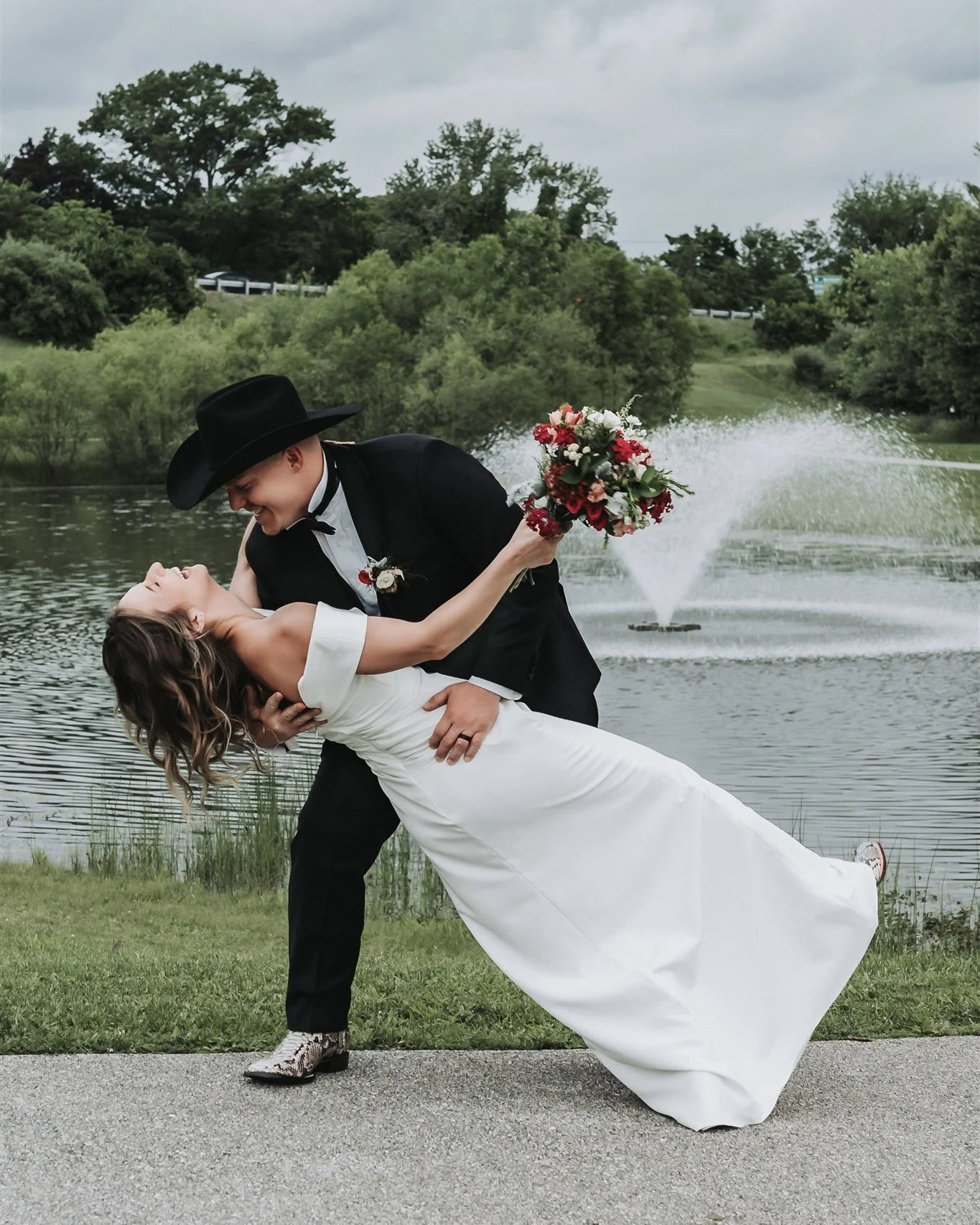 Groom in a black cowboy hat dips his laughing bride beside a park fountain at a Maryland wedding