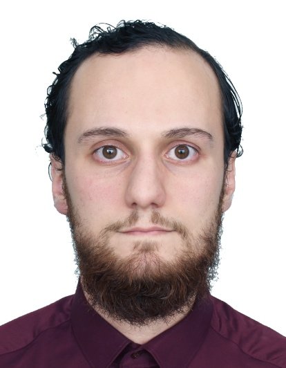 A man with dark, curly hair, a beard, and mustache, wearing a maroon shirt, looking directly at the camera with a neutral expression.