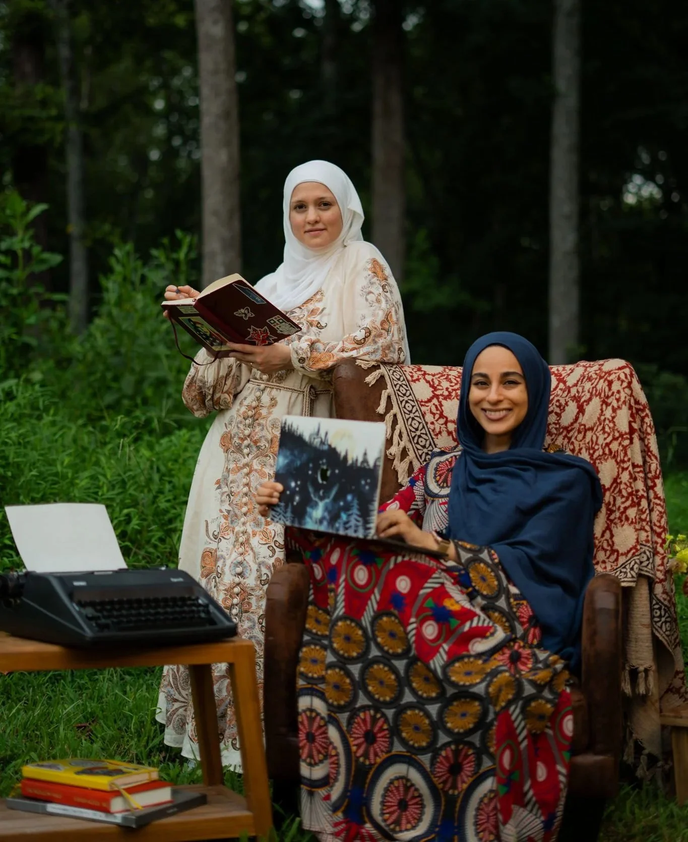 Two women wearing hijabs, one seated on a wooden chair with a colorful patterned skirt and blue hijab holding a book, and the other standing behind her with a white hijab, a book in hand, in a green outdoor forest setting.
