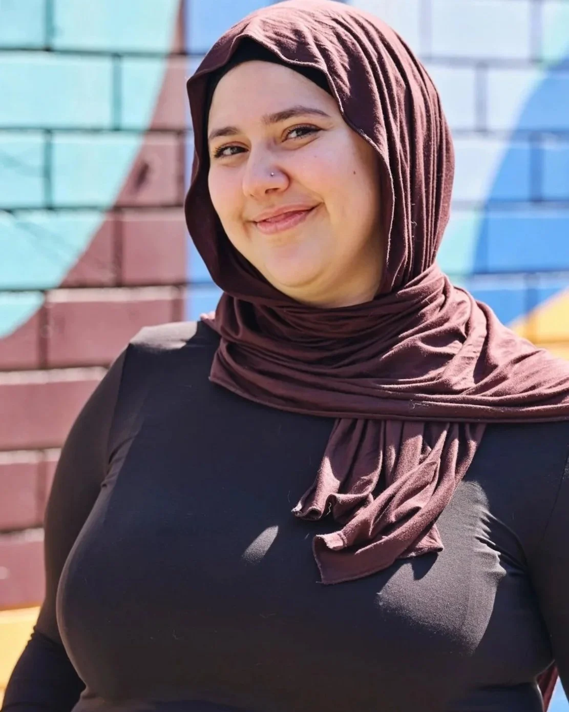 A young woman wearing a maroon hijab and black top, smiling, standing in front of a colorful painted brick wall.
