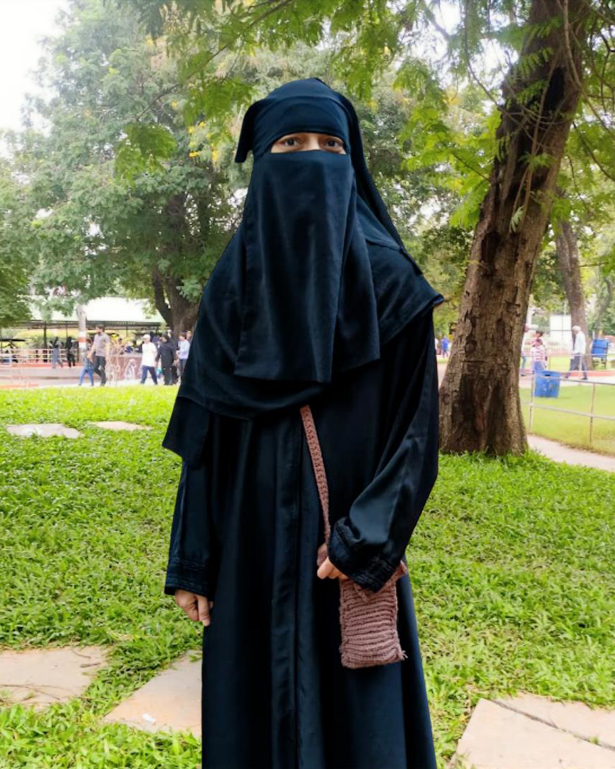 A woman is standing outdoors in a park, wearing a black hijab and niqab. She has a small brown bag hanging from her shoulder, and there are trees and people in the background.
