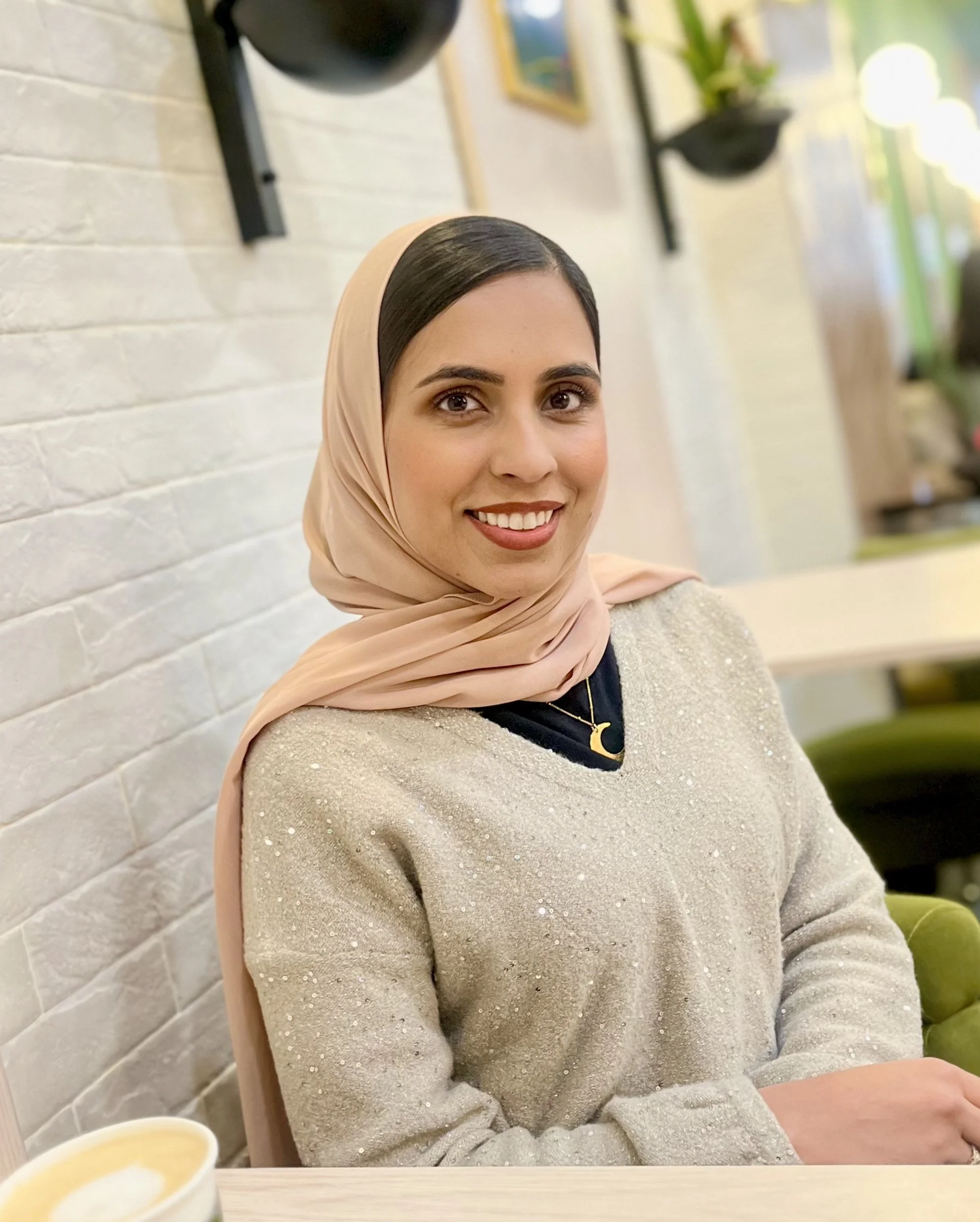 A woman with dark hair, wearing a light beige hijab, a sparkly beige sweater, and a gold crescent moon necklace, smiling while sitting in a cozy cafe with white brick walls and potted plants.