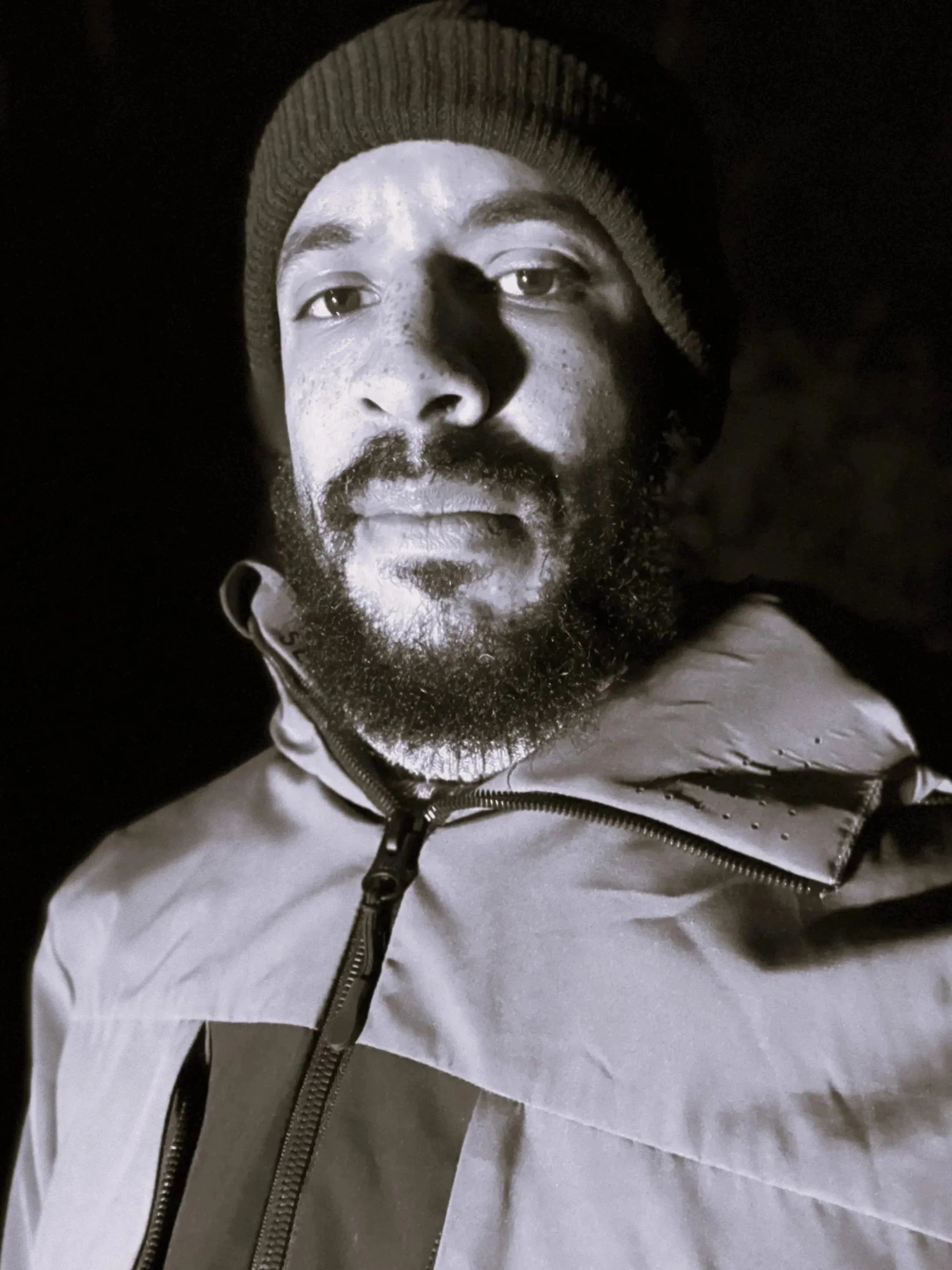 Black-and-white portrait of a man wearing a beanie and a zipped jacket, looking directly at the camera.