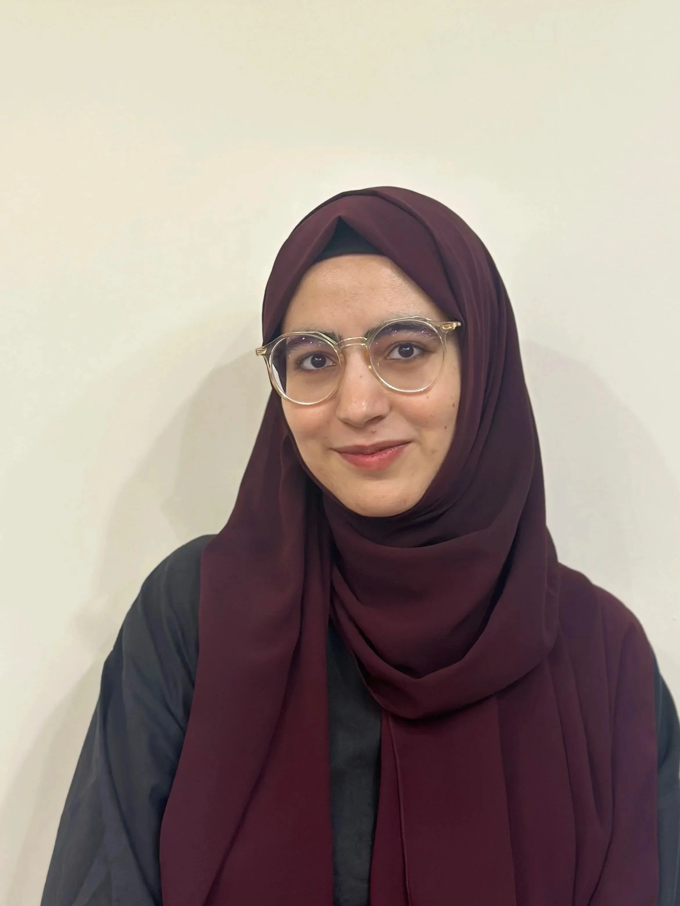 A woman wearing a maroon hijab and glasses, smiling against a plain light-colored wall.