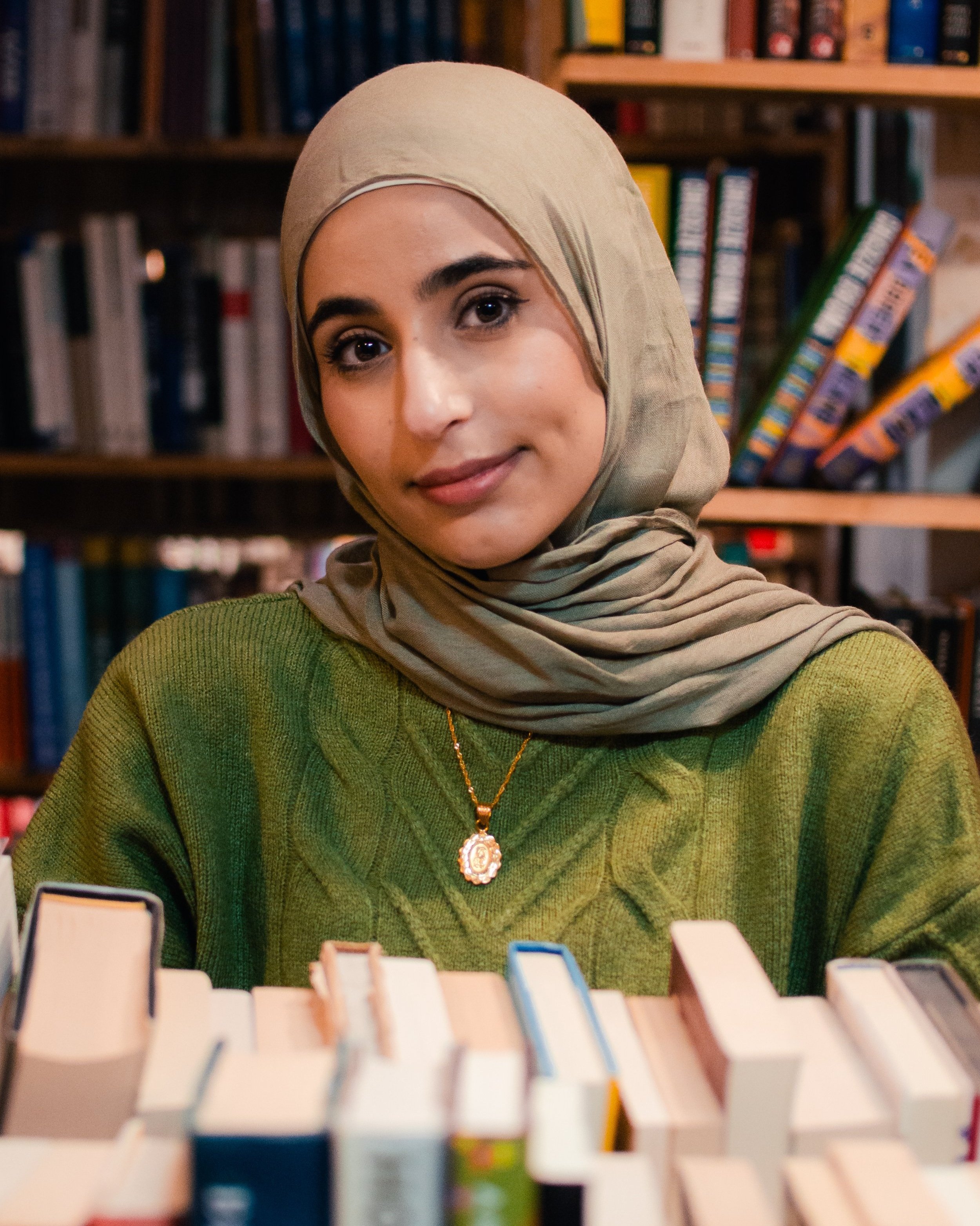 A young woman wearing a beige hijab and green sweater with a gold necklace, sitting in a library with shelves of books behind her.