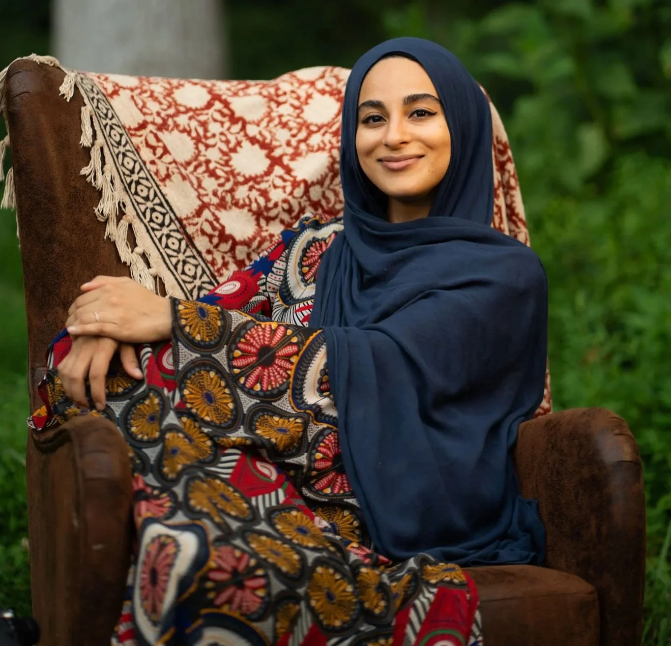 A woman in a colorful patterned dress and a navy blue hijab sitting on a brown armchair outdoors with greenery in the background, smiling at the camera.