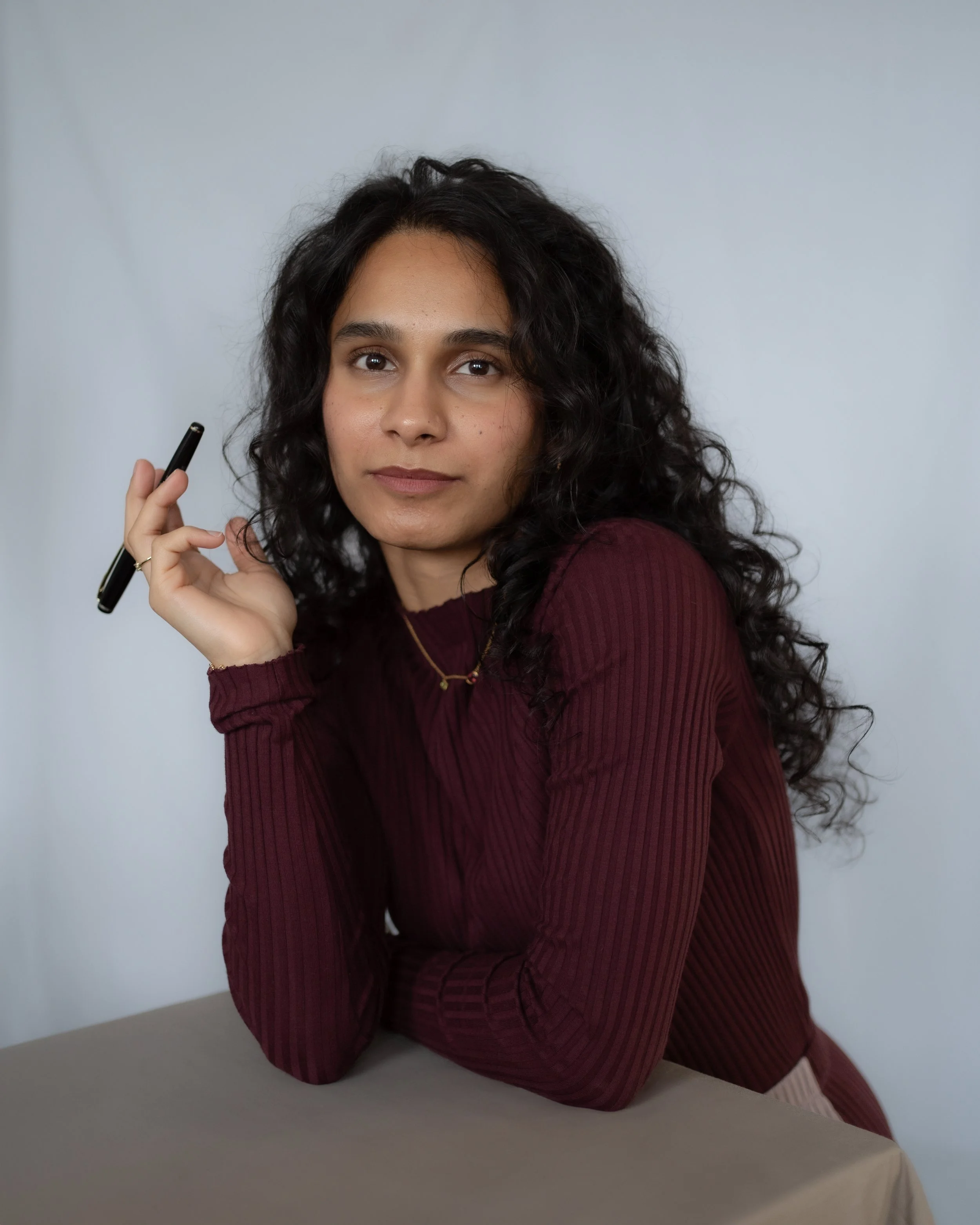 A woman with curly black hair, wearing a burgundy ribbed long sleeve shirt, holding a black pen in her right hand, sitting at a table with a neutral expression, against a plain light gray background.