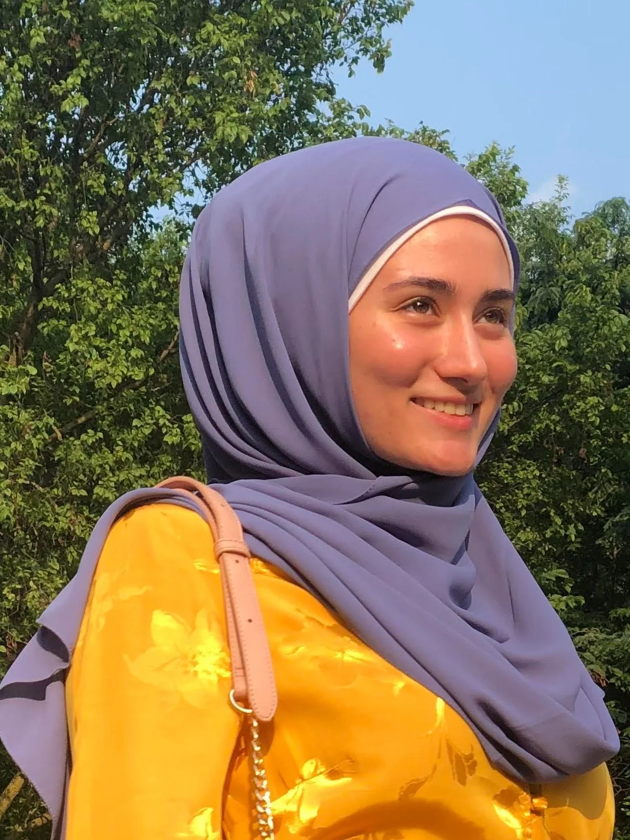 A young woman wearing a purple hijab and a shiny yellow top, standing outdoors with green trees and a blue sky in the background.