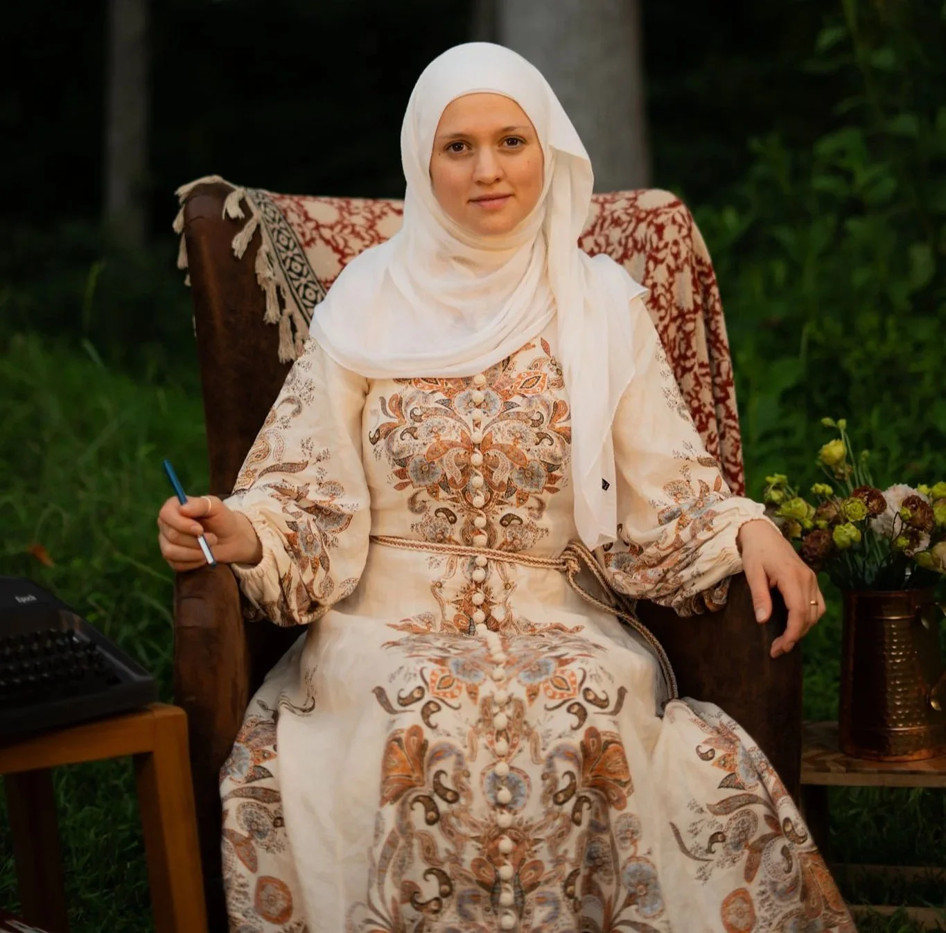 Woman in traditional embroidered dress and white headscarf sitting on a chair outdoors, holding a pen, with a small wooden table and a potted plant nearby.