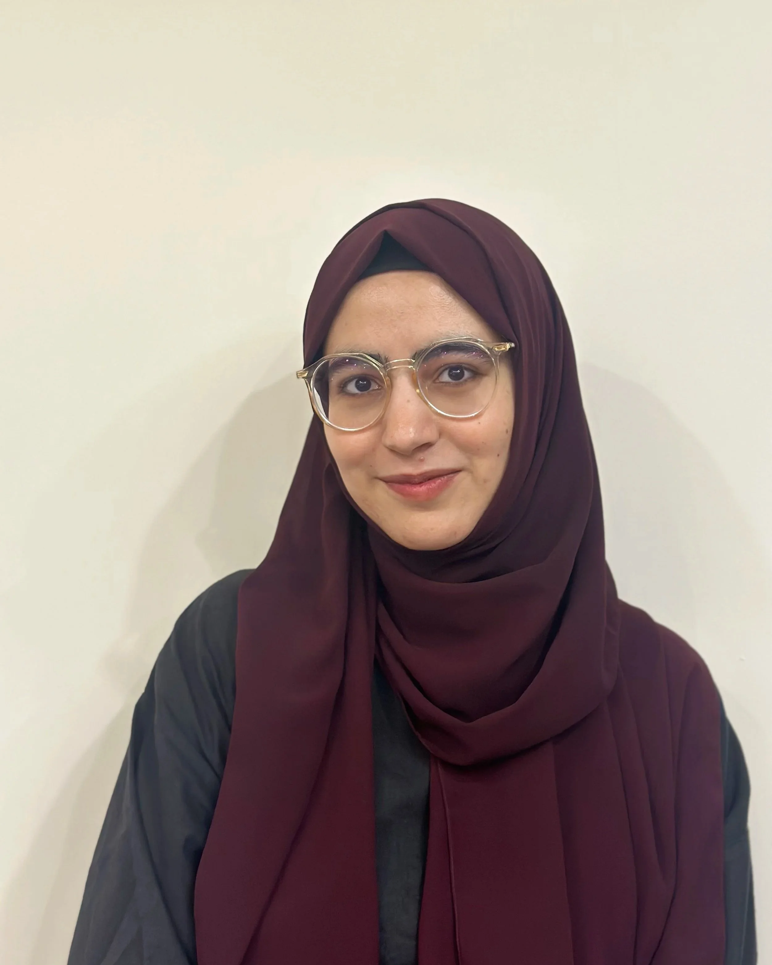 A woman wearing glasses and a maroon hijab, smiling softly, standing against a plain white wall.