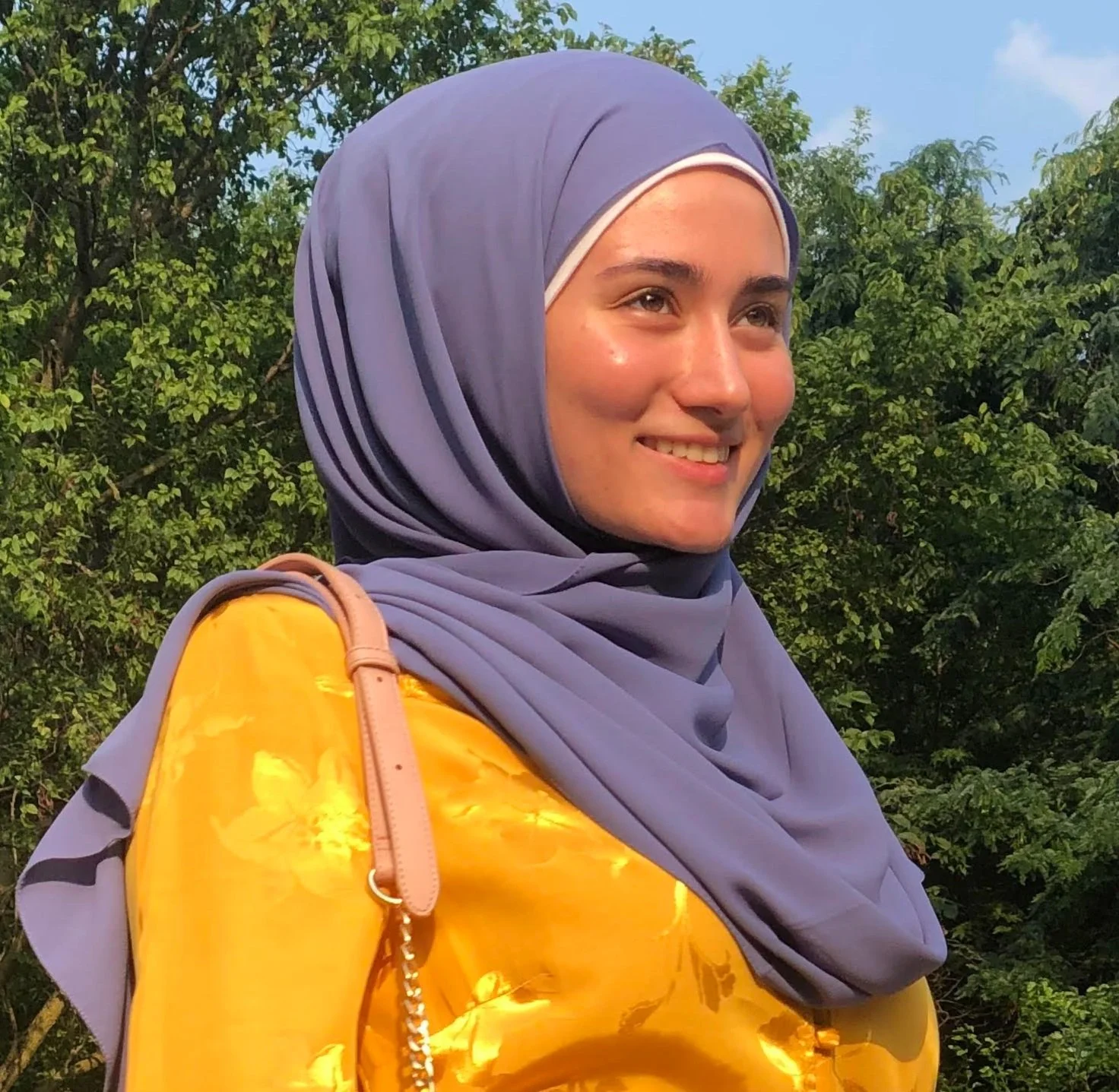 A young woman wearing a lavender hijab and a yellow jacket, smiling outdoors with green trees in the background.
