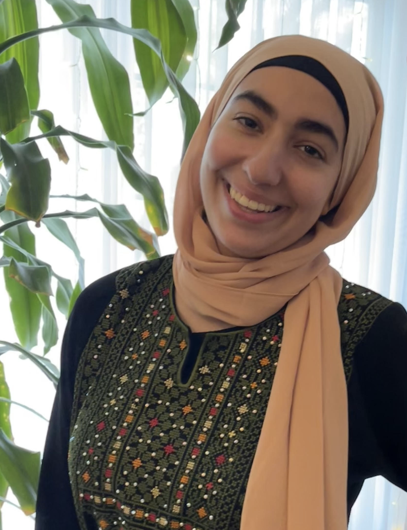 Young woman wearing a beige hijab smiling, standing in front of a tall green plant and white curtains.