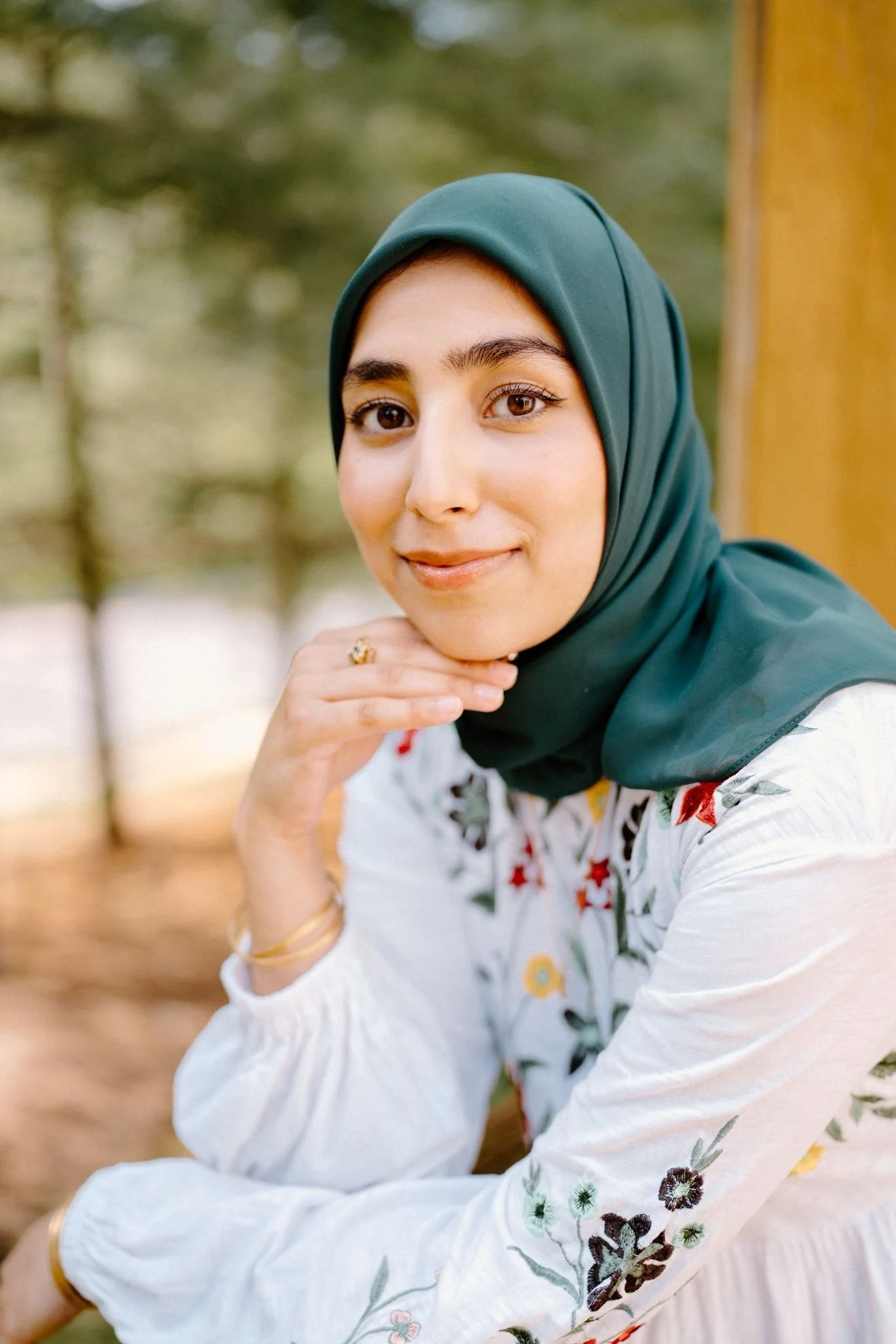 A woman wearing a teal hijab and a white embroidered blouse, sitting outdoors with trees in the background, smiling and resting her chin on her hand.