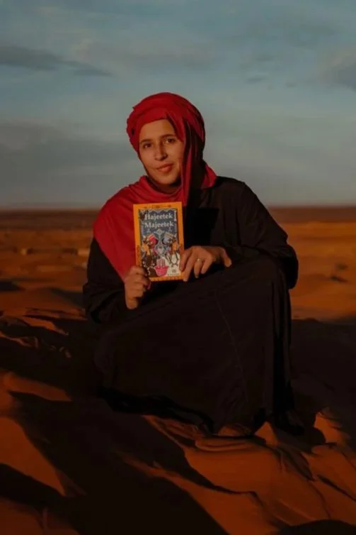 A young person dressed in black sitting on desert sand dunes holding a book with a blue cover.