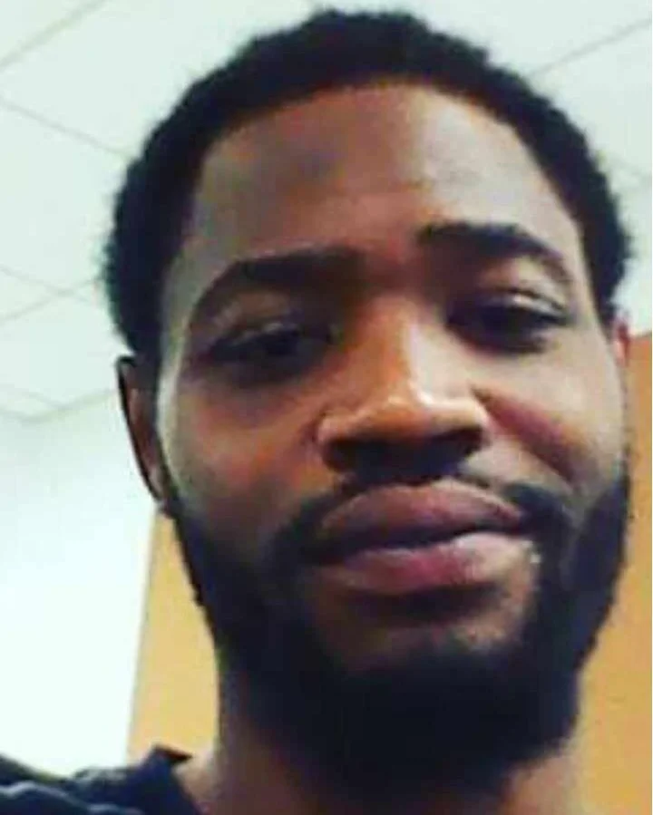 Close-up of a young man with dark skin, short curly hair, and a beard, smiling in an indoor setting.