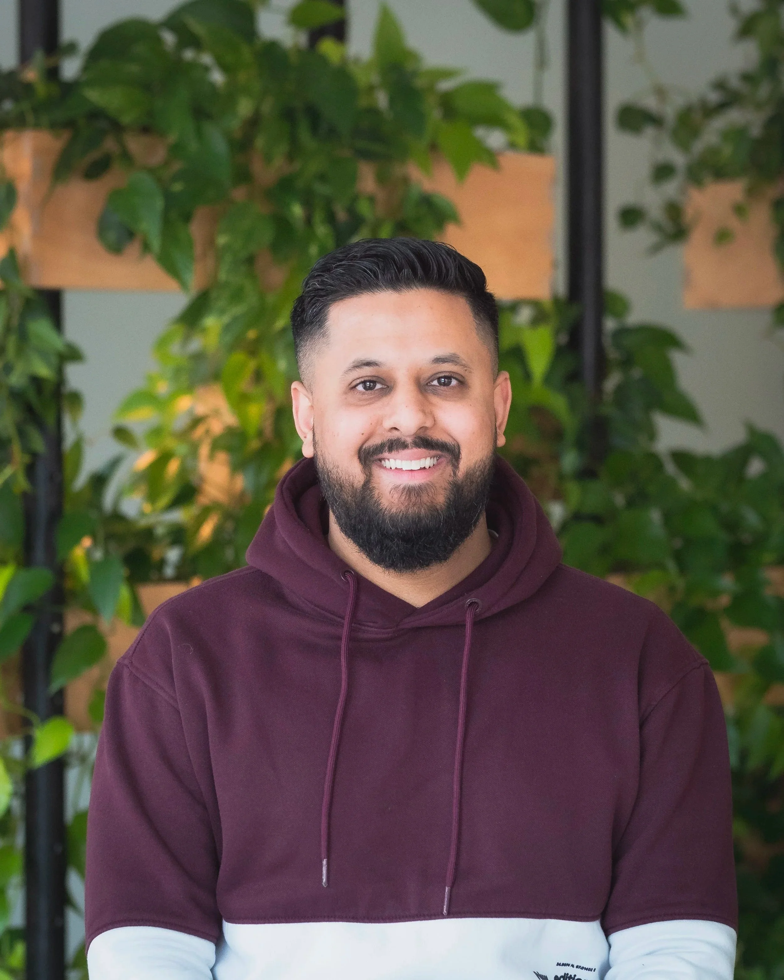 A man with a beard and short dark hair smiling in front of green leafy plants indoors.