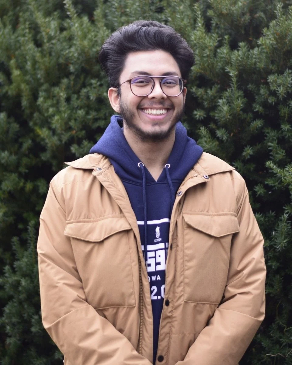 A smiling young man with glasses, wearing a tan jacket and a navy hoodie, standing outdoors in front of green shrubbery.