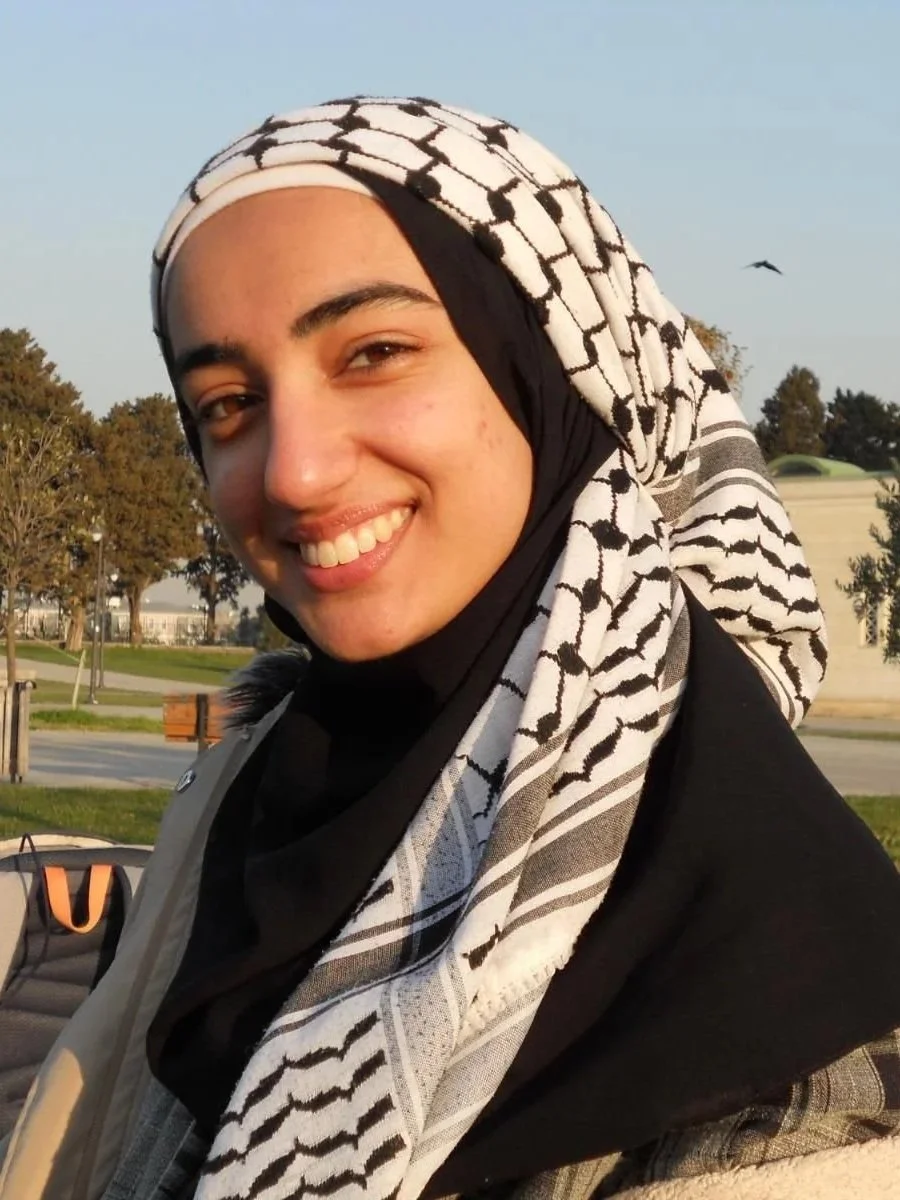A young woman wearing a black and white keffiyeh scarf smiling outdoors in a park with trees and a bird flying in the background.