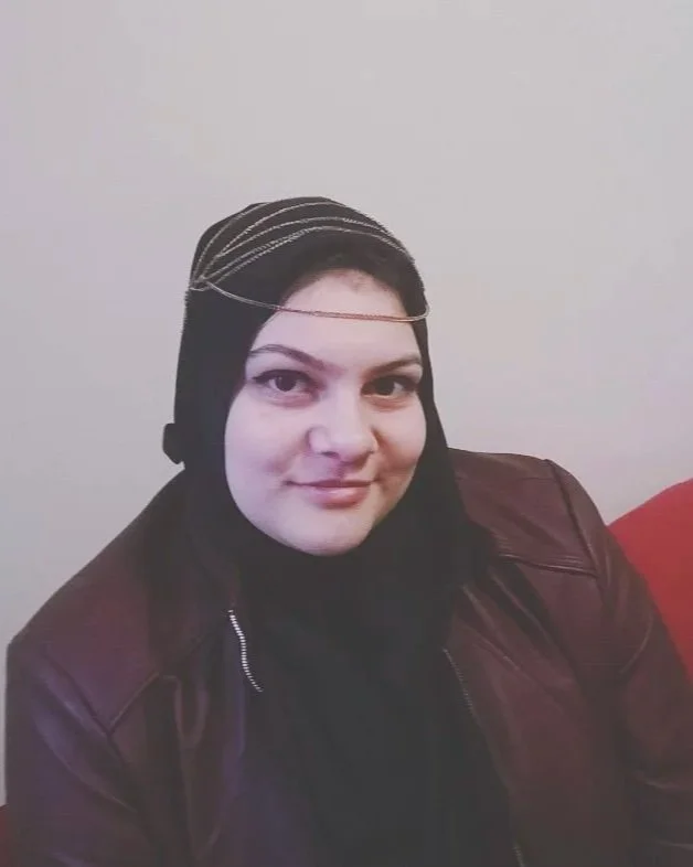 A woman wearing a black hijab and a dark brown leather jacket, sitting on a red couch against a light-colored wall.