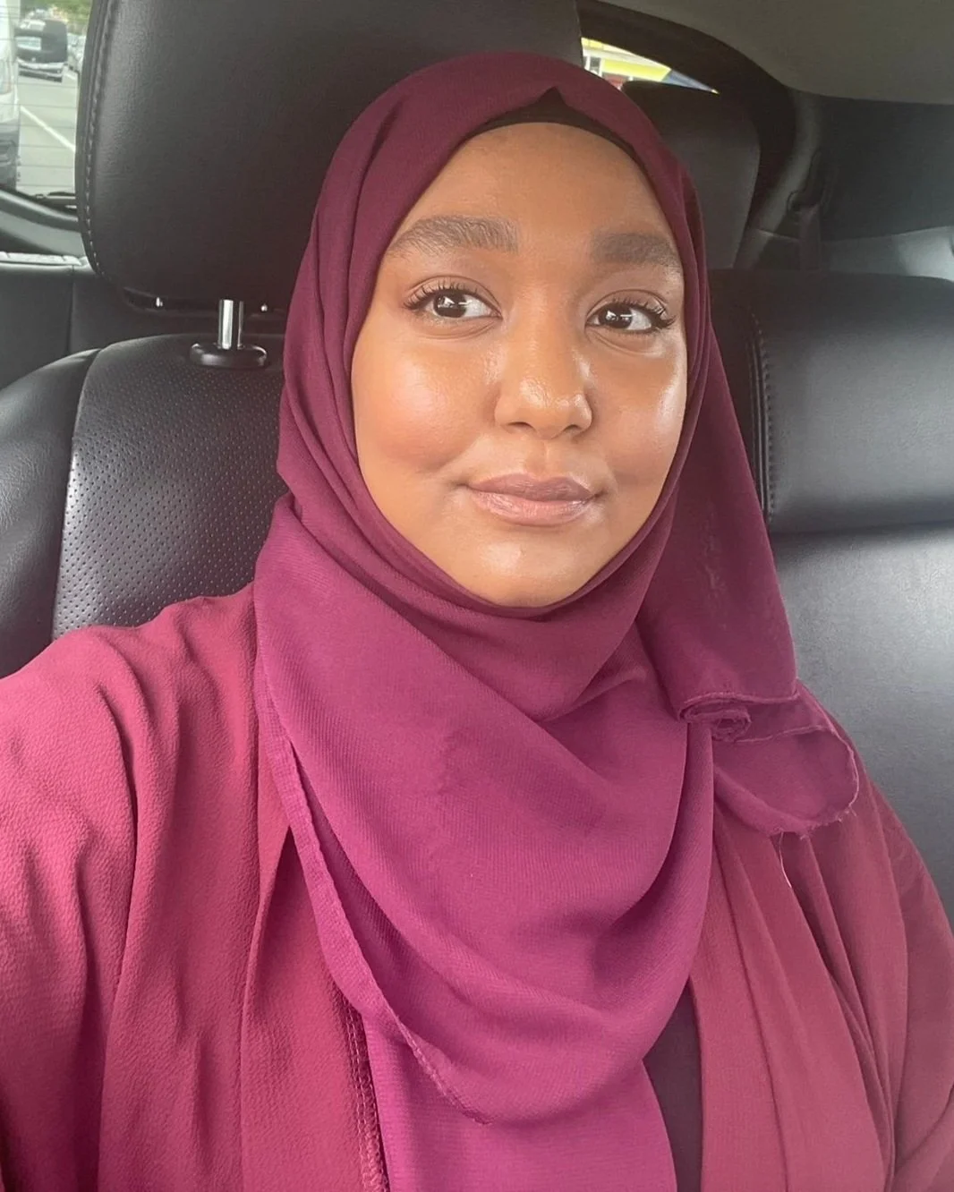 A woman in a maroon hijab and matching top sitting in a car seat inside a vehicle.