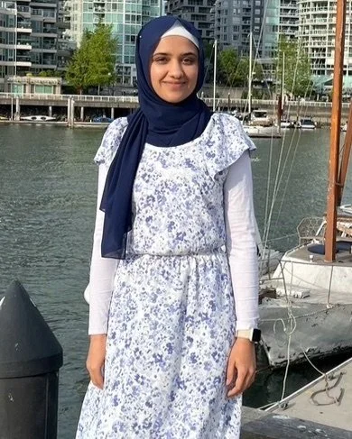 A woman wearing a blue headscarf and a white and blue floral dress standing by a marina with boats and modern buildings in the background.