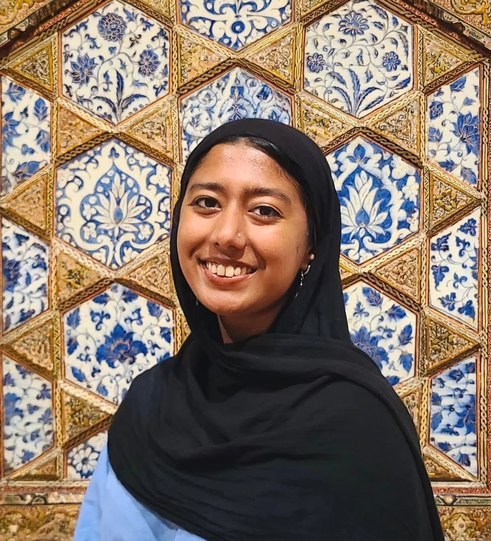 A young woman wearing a black headscarf and light blue top, smiling in front of an ornate blue and gold tile wall with intricate floral and geometric patterns.