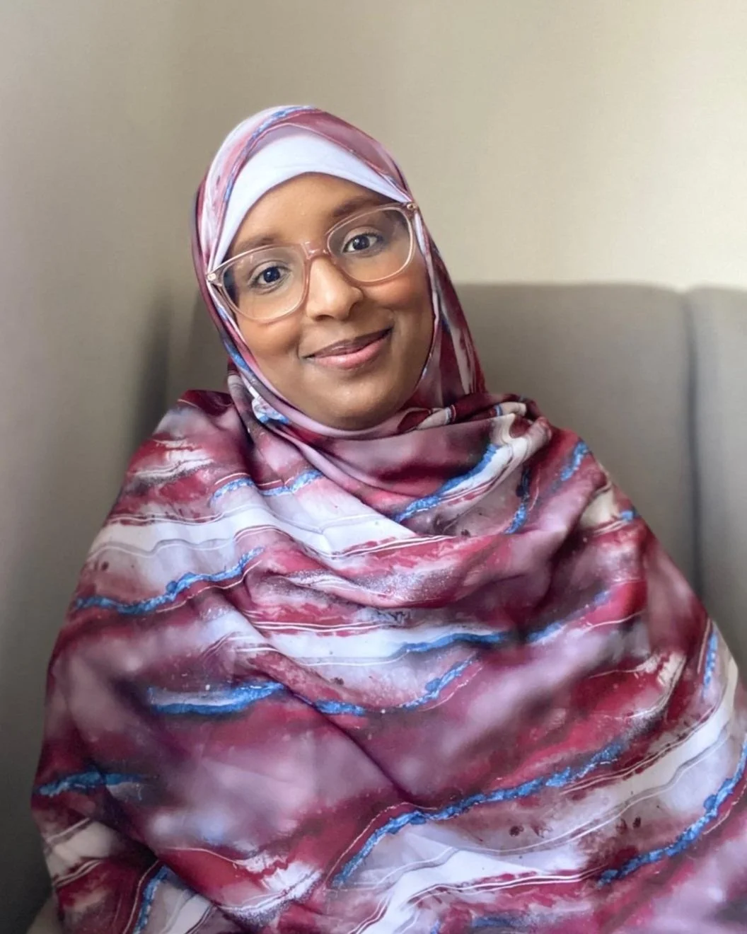A woman smiling, wearing glasses and a colorful, patterned hijab, sitting against a plain, light-colored wall.