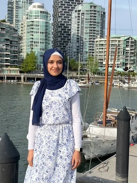A woman in a blue hijab and white floral dress standing on a marina dock with boats, with city buildings in the background.