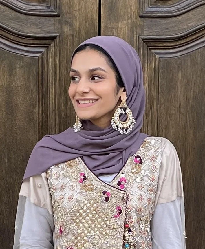 A woman wearing a lavender headscarf, large pearl and gold earrings, and an ornate cream-colored top with embroidery and pink accents, standing in front of a wooden door.