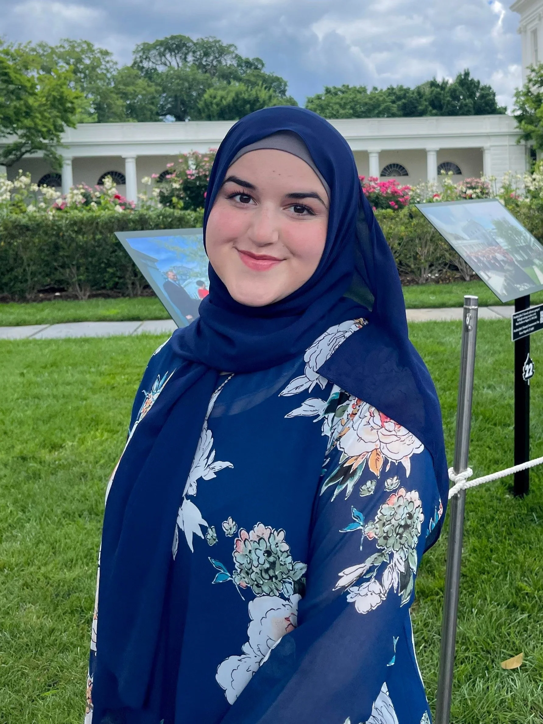 A young woman wearing a dark blue floral hijab and matching floral dress, smiling outdoors in front of a white building with columns, greenery, and display boards.