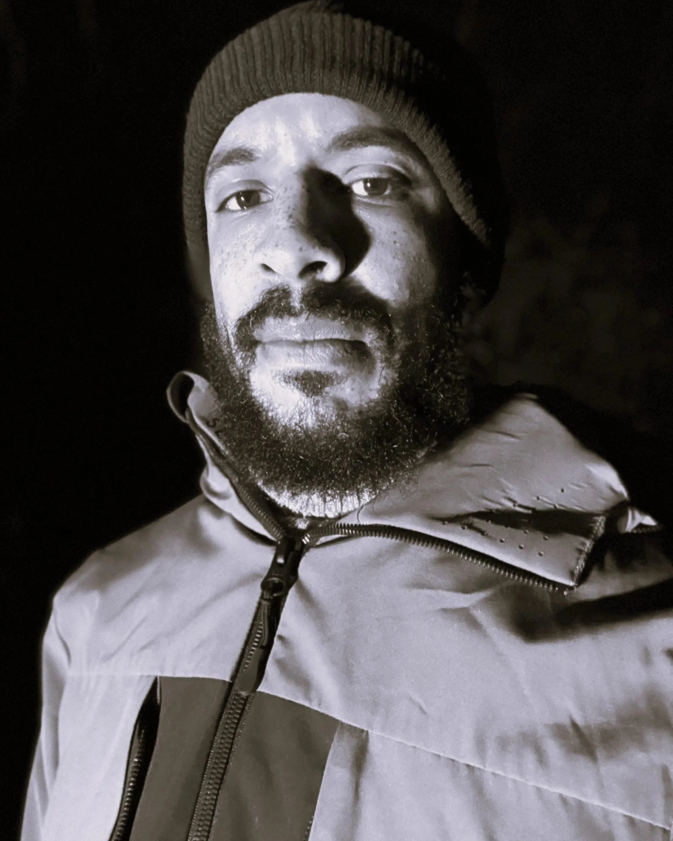 Black and white close-up photo of a man wearing a beanie and a jacket, looking directly at the camera.