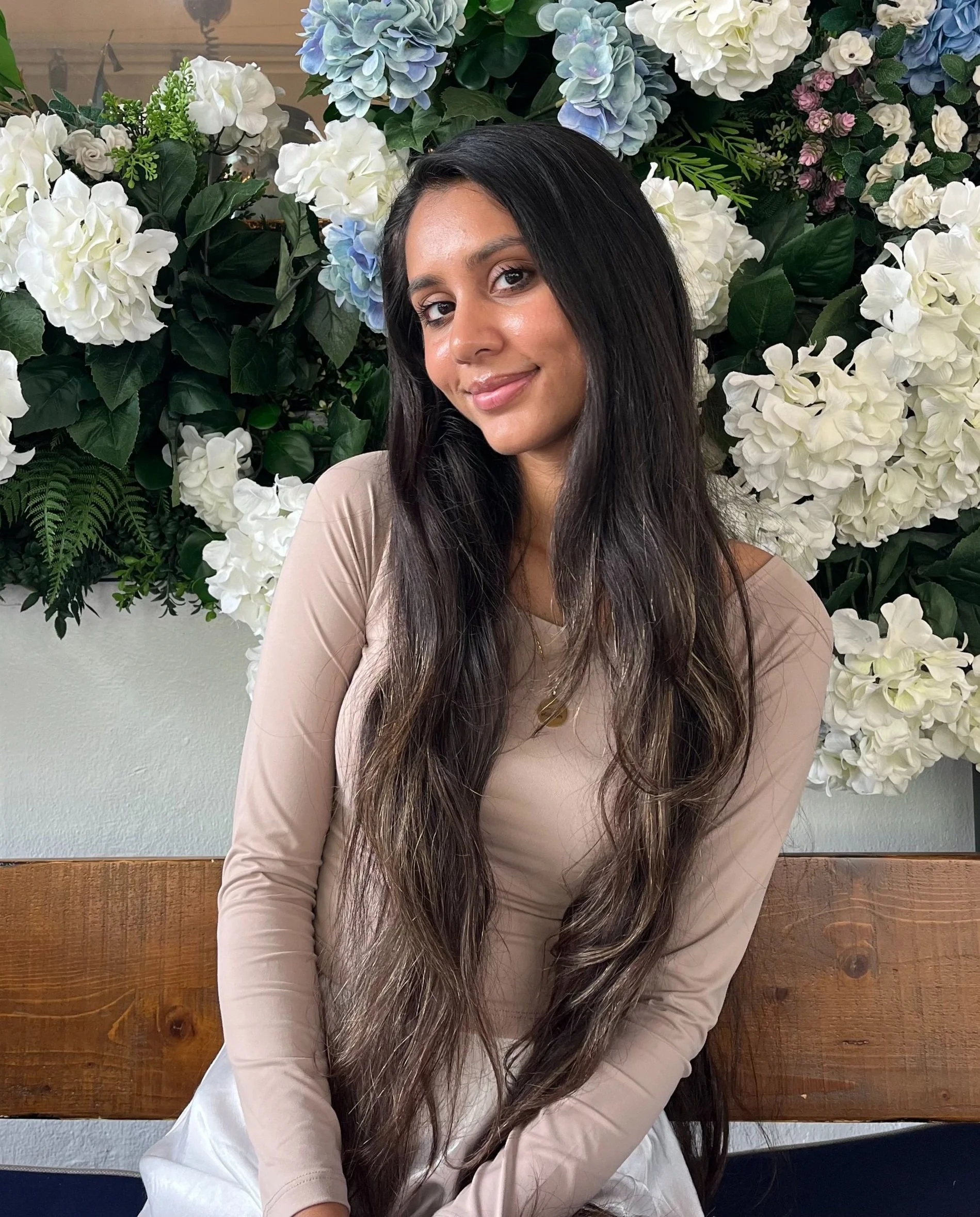 A young woman with long, dark wavy hair and a tan complexion smiling while sitting on a wooden bench against a backdrop of white and purple flowers, including hydrangeas.