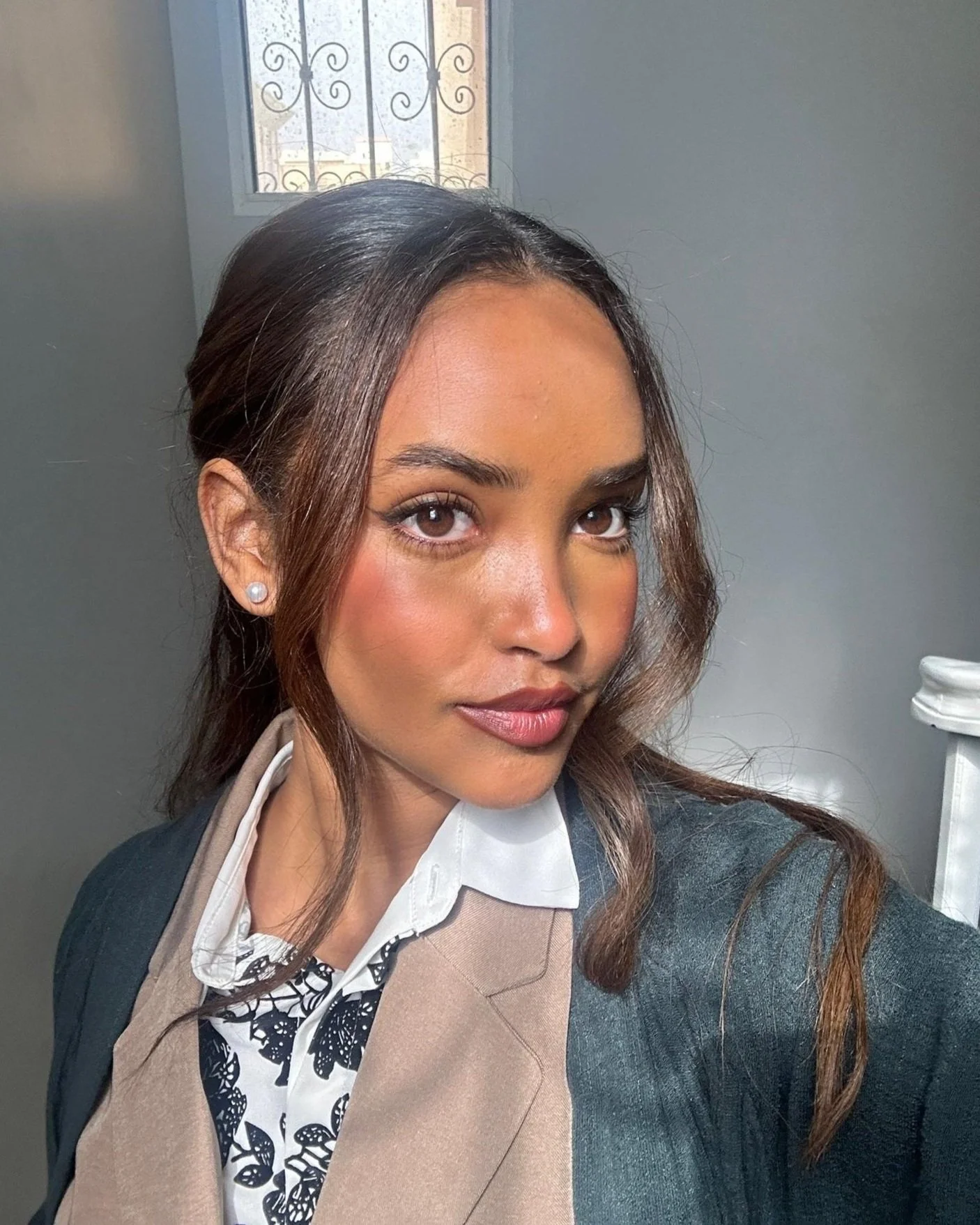 A woman with medium brown skin and brown hair styled in loose waves, wearing pearl earrings and a formal blazer over a printed shirt, looking at the camera with a neutral expression.