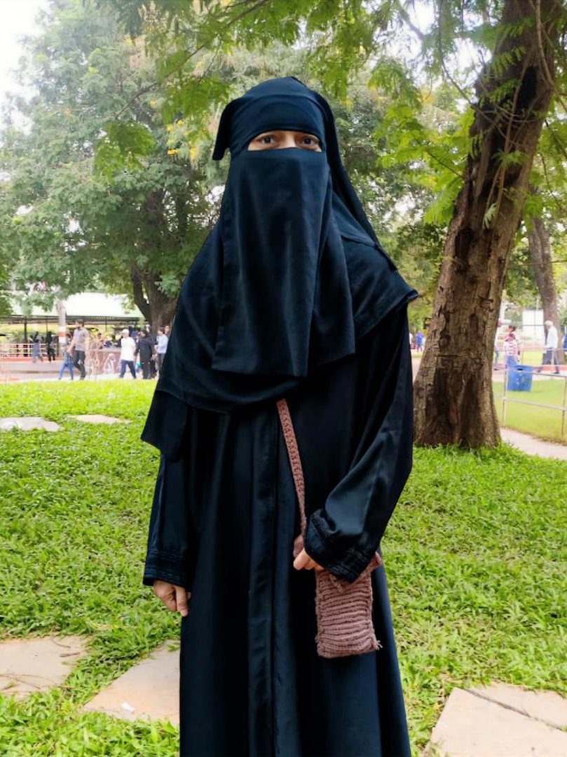 Woman dressed in a black abaya and niqab standing outdoors in a park with green grass and trees, with people visible in the background.