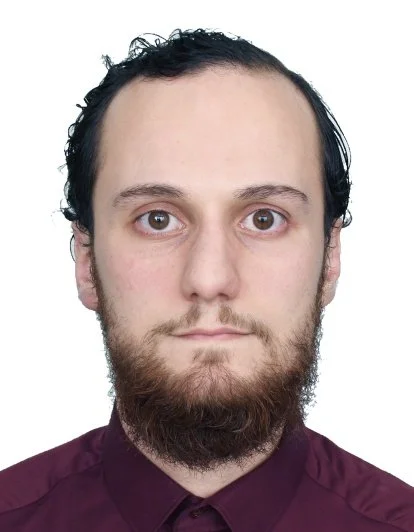 Headshot of a man with dark, curly hair, a beard, and light skin, wearing a maroon shirt against a white background.