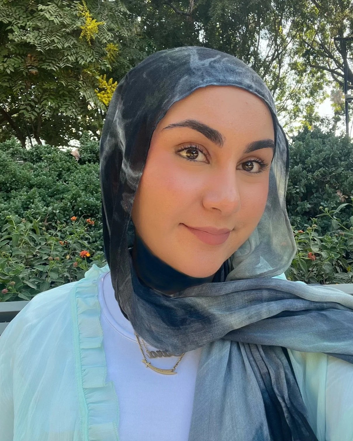 A woman wearing a black and gray scarf, a white top, and a gold necklace, standing outdoors in front of green bushes and trees, taking a selfie.