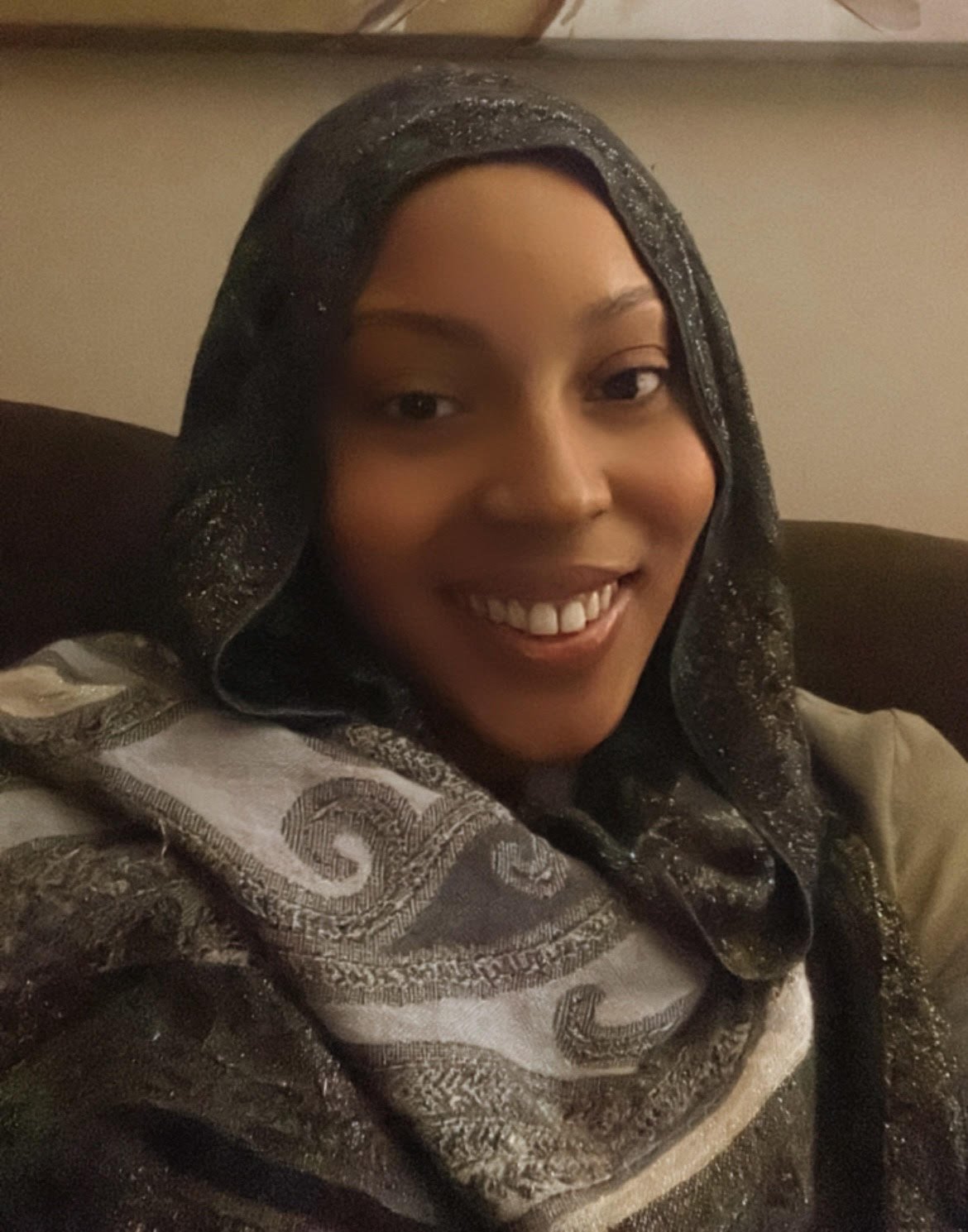 A woman smiling, seated on a dark-colored couch, wearing a dark scarf with sparkles and a patterned jacket.