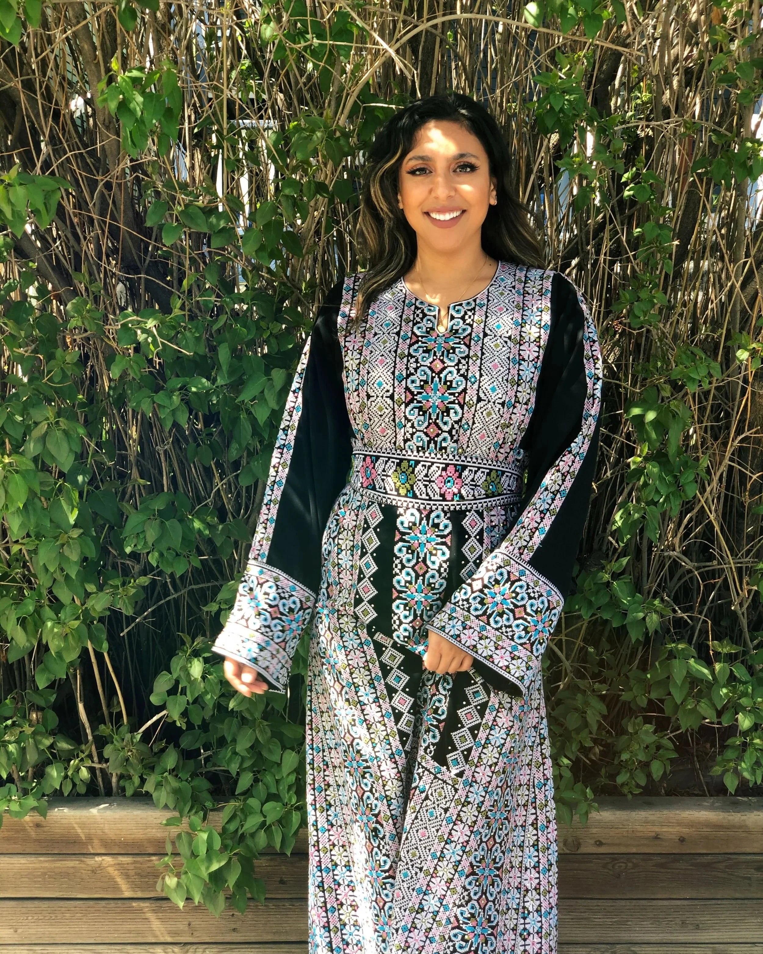 Woman wearing a traditional embroidered dress standing outdoors in front of green foliage.